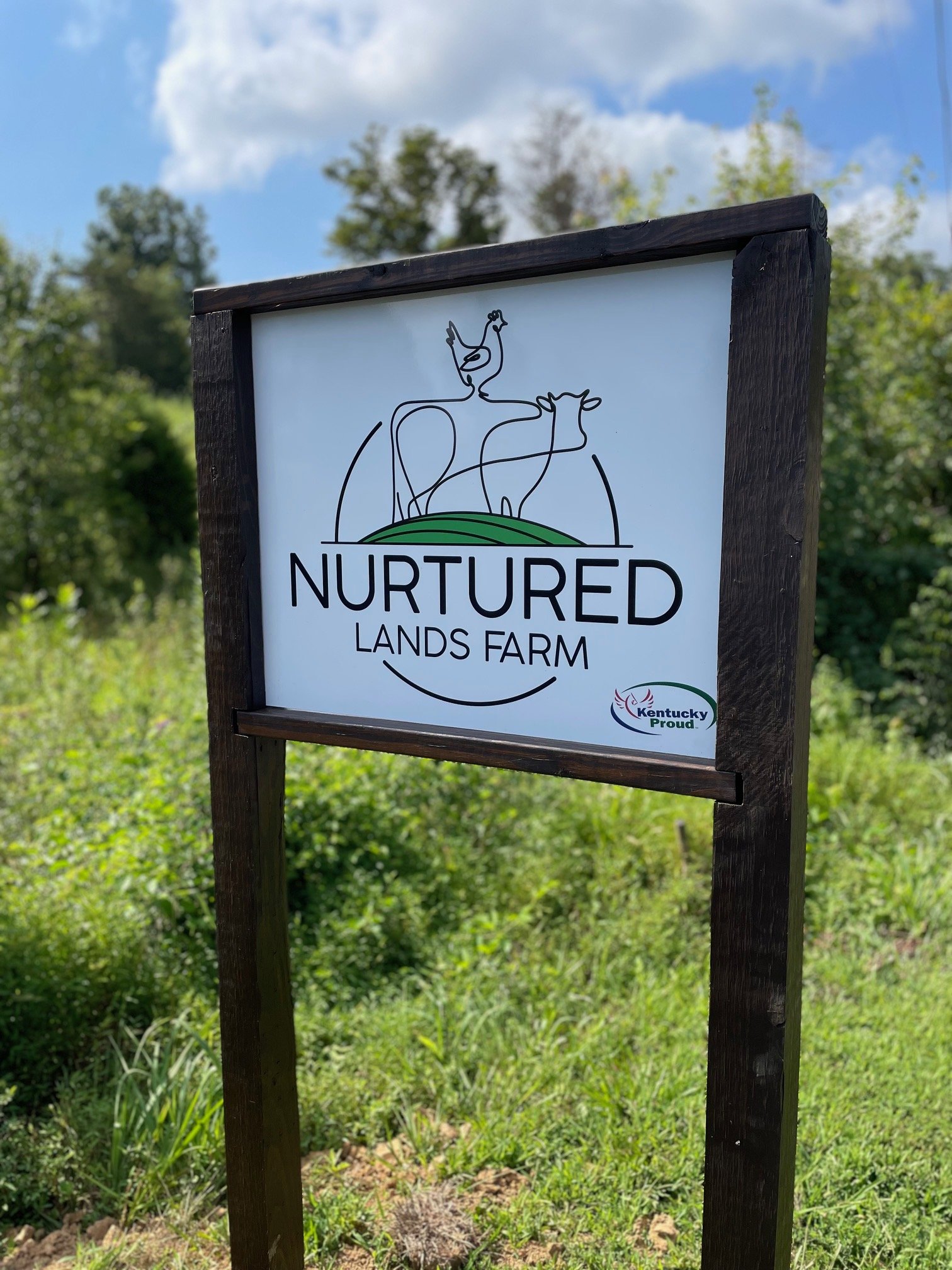 A sign outside with a simple line drawing of a rooster and cow above the words "Nurtured Land Farm" with a small logo of Kentucky Proud at the bottom right corner, against a backdrop of trees and a partly cloudy sky.