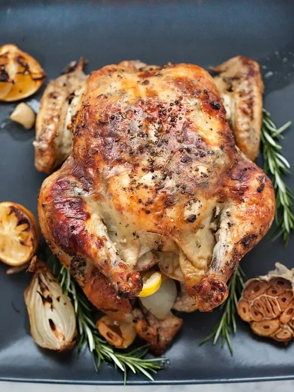 Roasted whole chicken garnished with herbs and surrounded by grilled vegetables on a black serving platter.