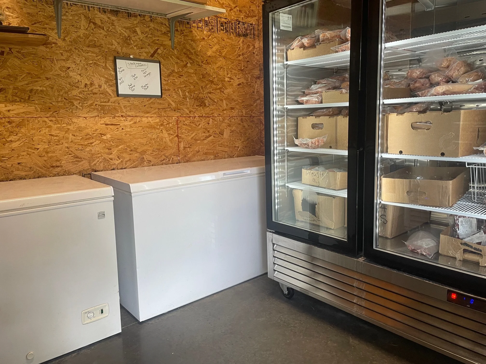 Frozen meats stored inside a glass-front refrigerator in a grocery or butcher shop, with cardboard boxes on the shelves and a wooden wall behind.