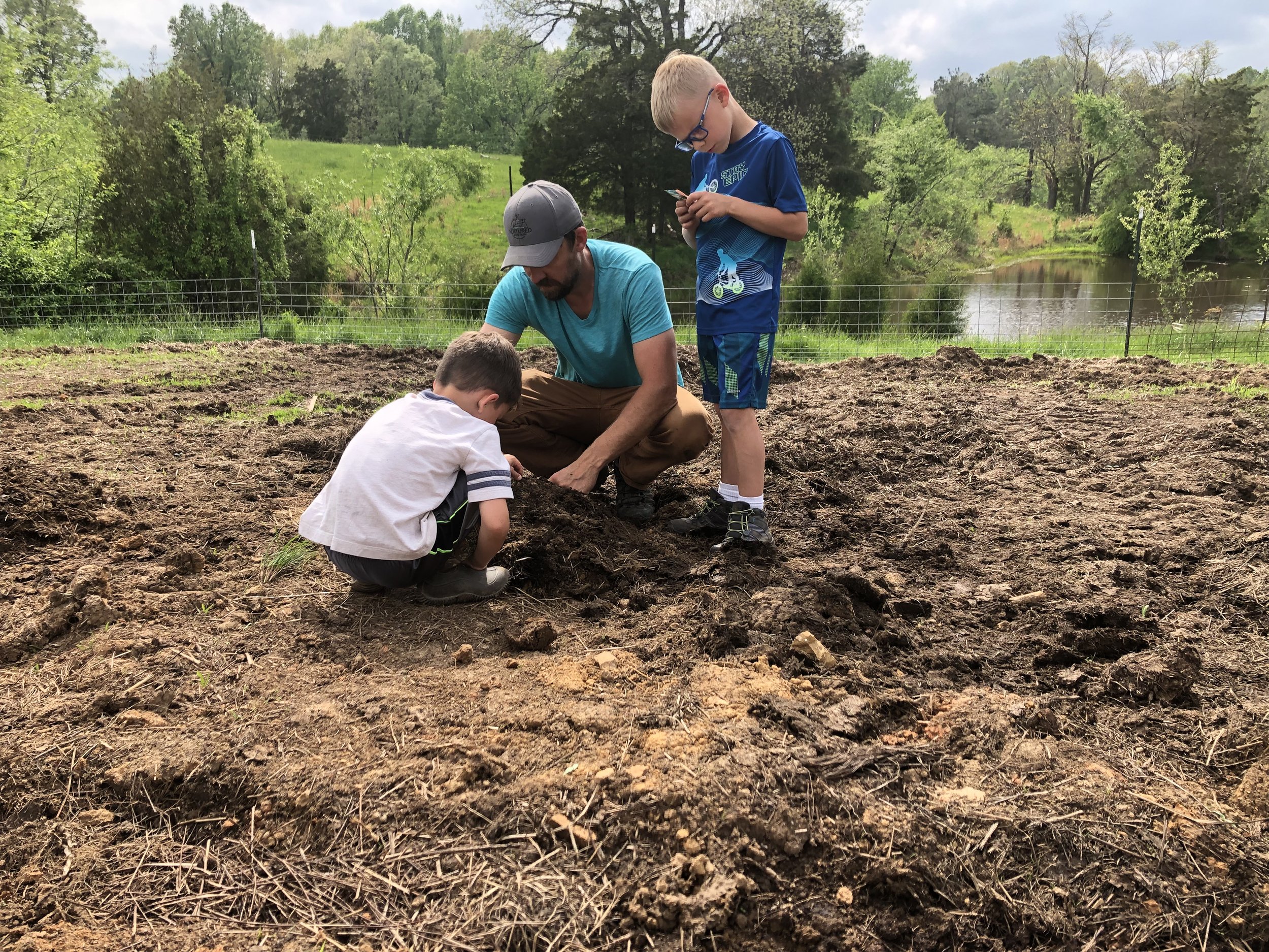 Two children and an adult planting a seed in a garden with bare soil, a fence, trees, and a pond in the background.
