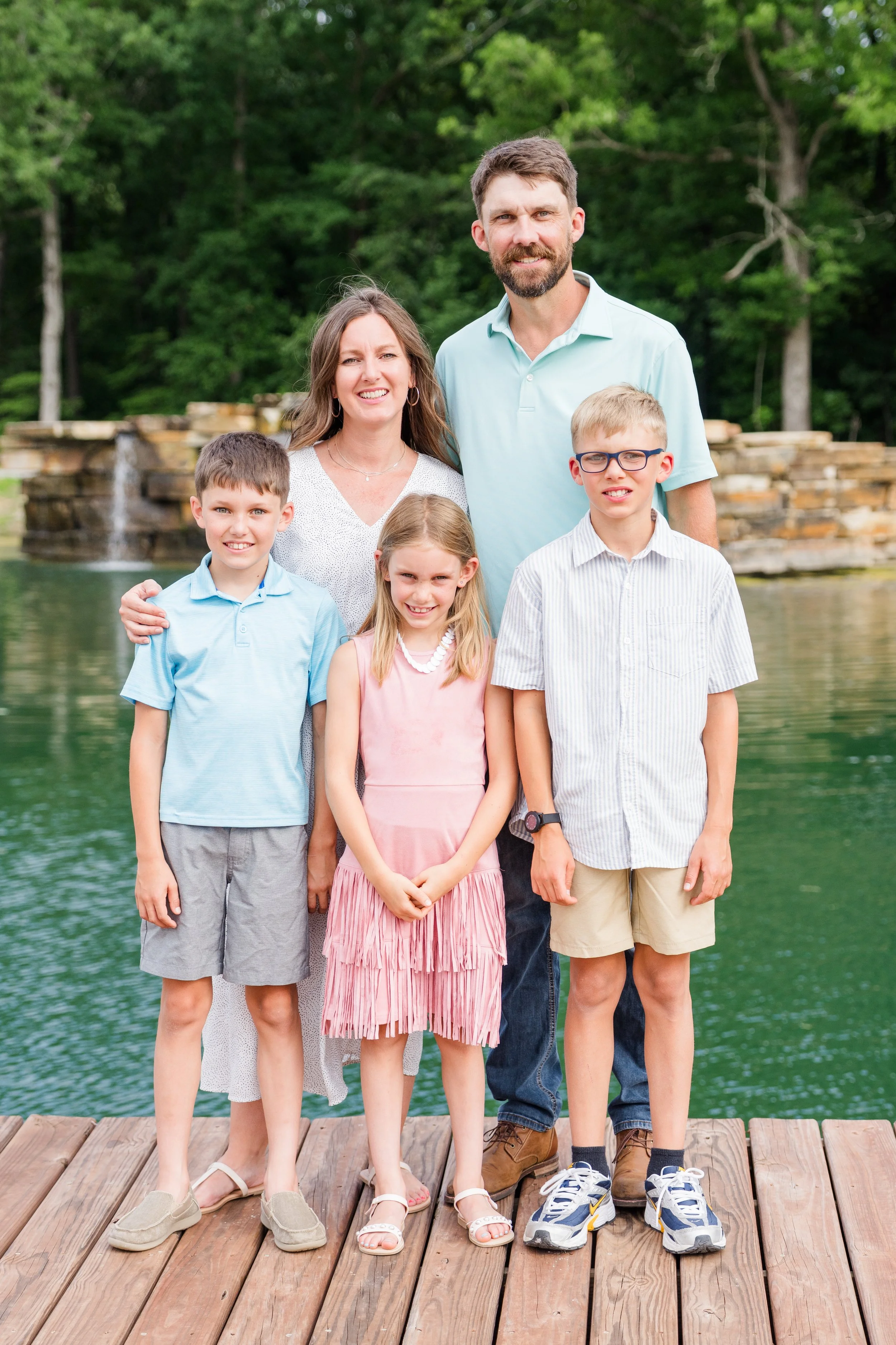 Family of six standing on a wooden dock near a body of water with trees in the background. Two adults and four children smiling at the camera.
