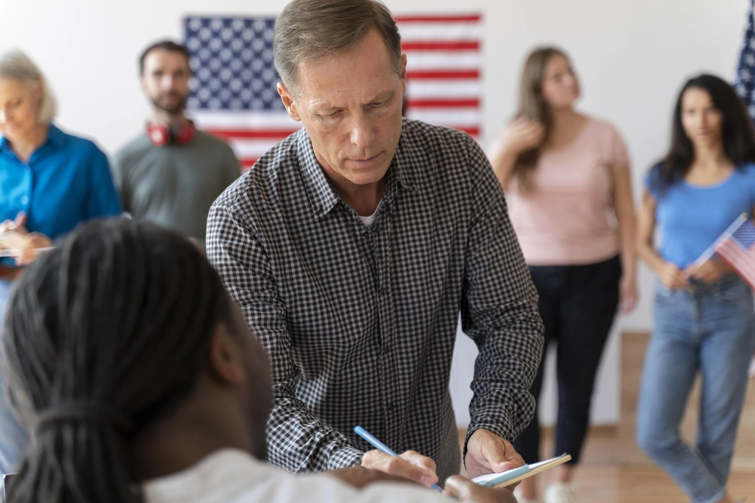A man in a checkered shirt is writing on a notepad while speaking with a young girl in a white top, in a room decorated with American flags; other people are in the background.