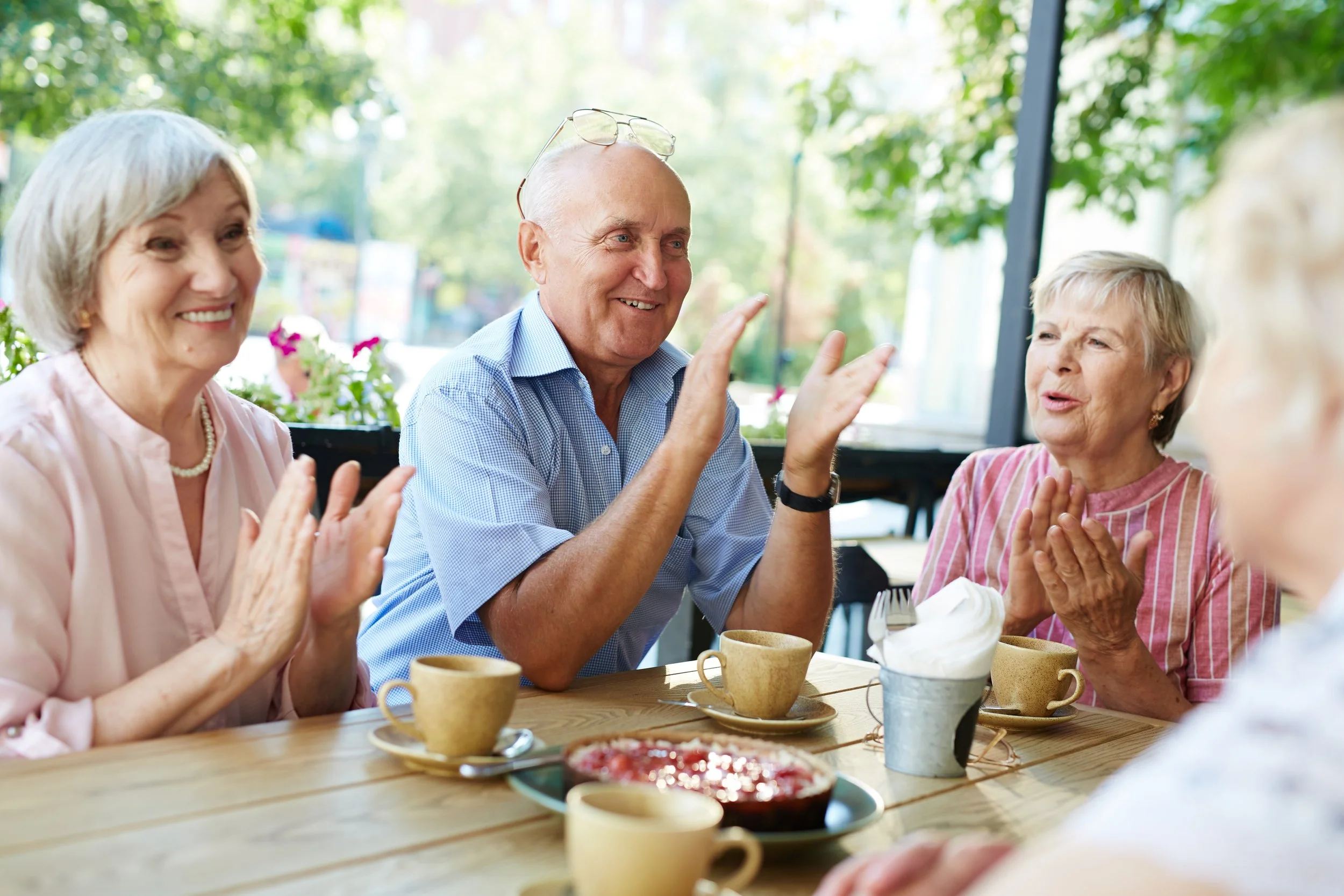 Four elderly women sitting at a table outdoors, clapping and smiling during a meal with cups, bowls, and food on the table.