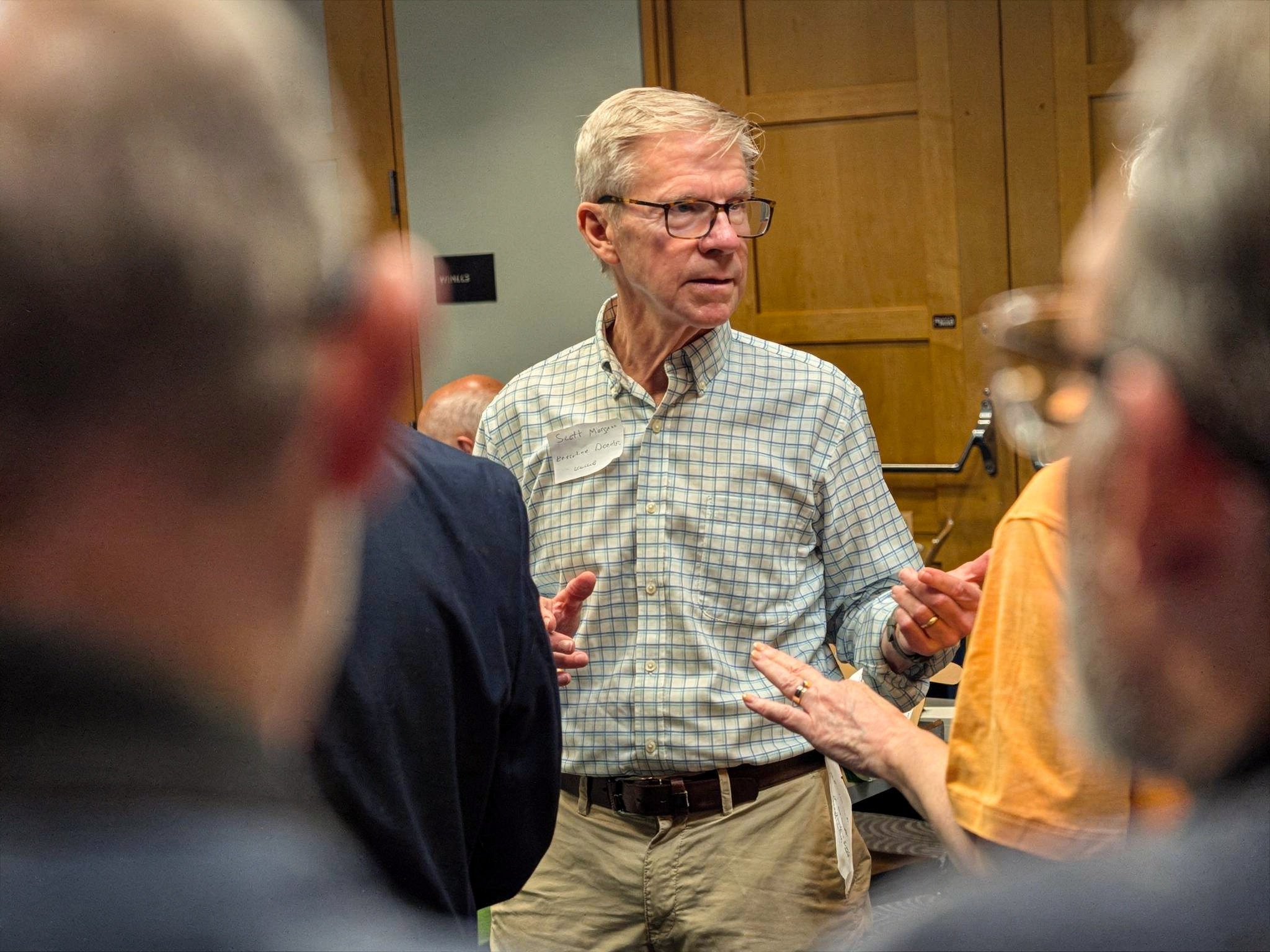 A man with gray hair, glasses, and a checkered shirt engaged in conversation at an indoor event, surrounded by other people.