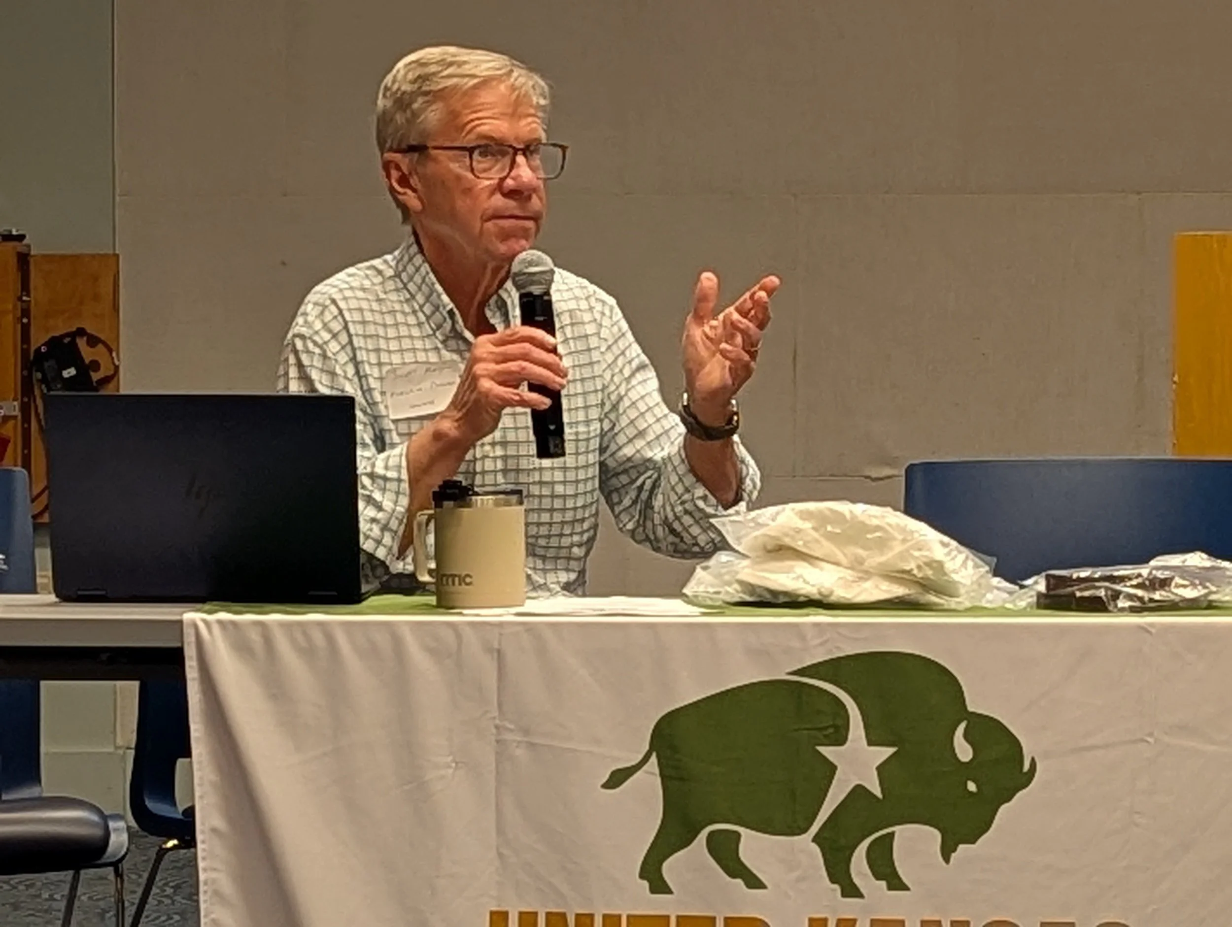 A man with glasses speaking into a microphone at a table with a laptop, coffee mug, and some bags. Behind him is a blank wall.
