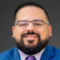 A man with glasses, a beard, and a mustache is smiling, wearing a blue suit and a purple tie.