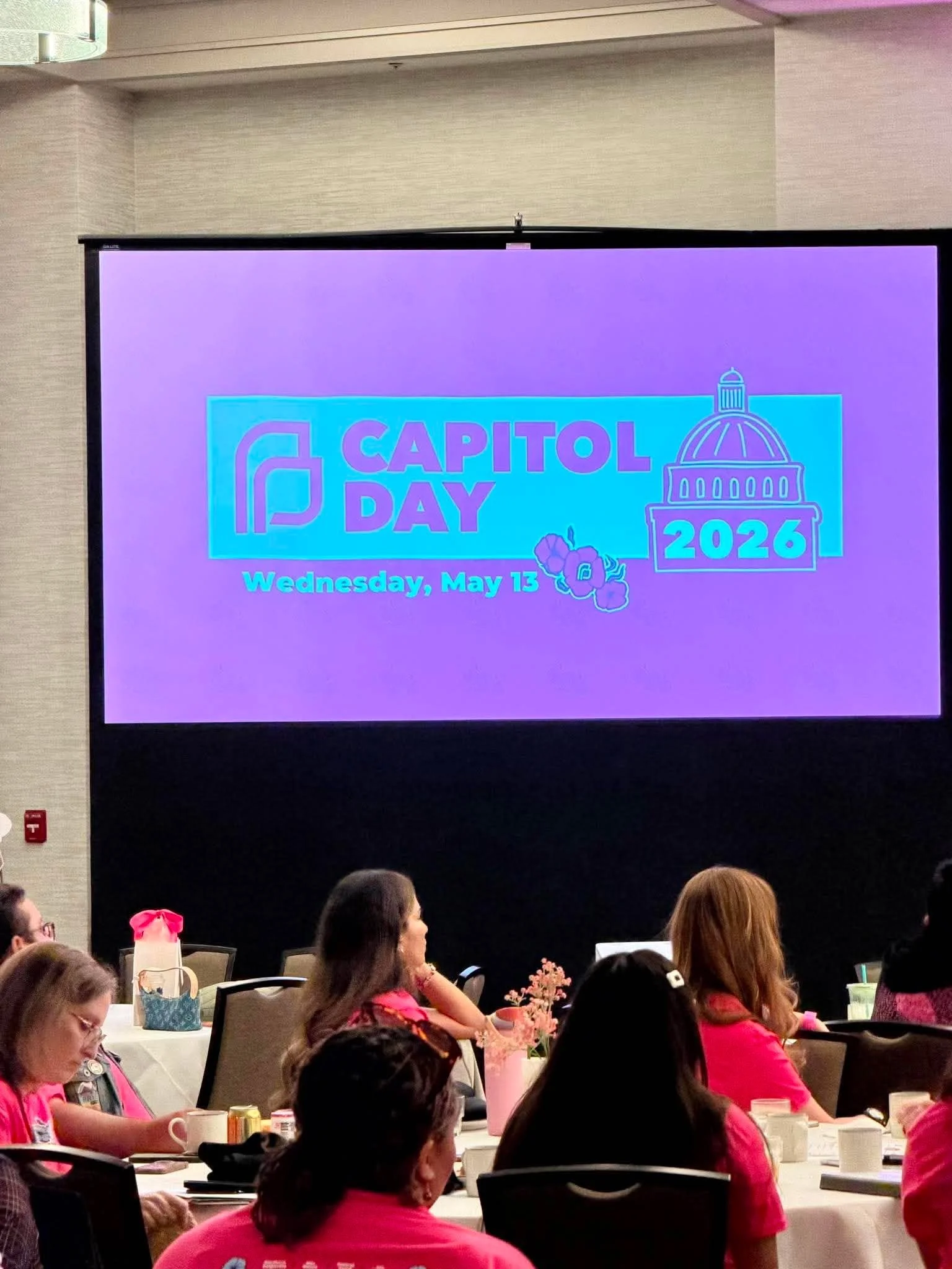 A large screen displaying a Capitol Day 2026 event announcement, with a purple background, event date Wednesday, May 13, and illustrations of the Capitol building, hibiscus flowers, and a person. Attendees seated at round tables in a conference room.