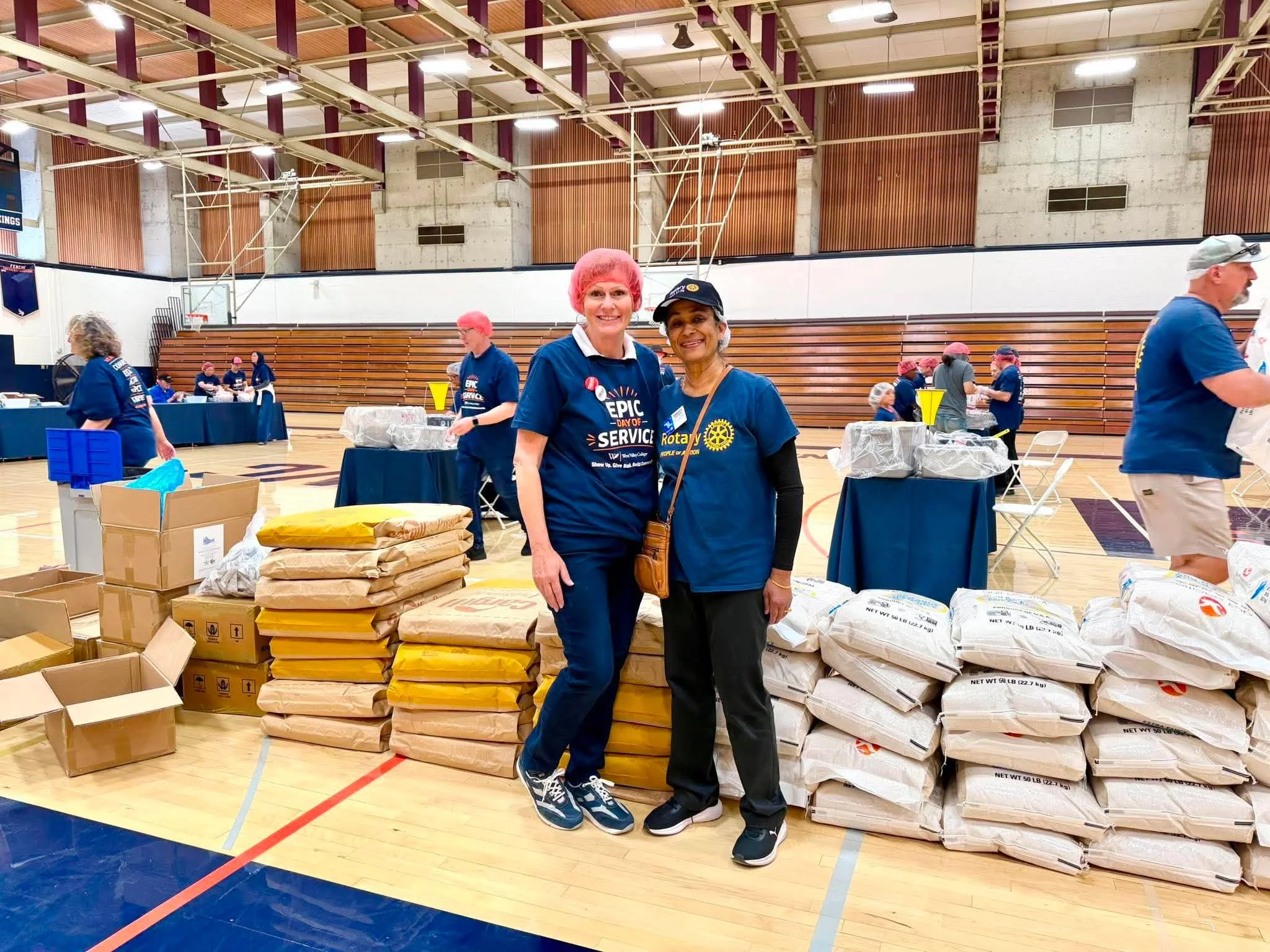 People at a charity event in a gymnasium, wearing blue shirts with 'Day of Service' and Rotary logos, surrounded by boxes of supplies and food packages.