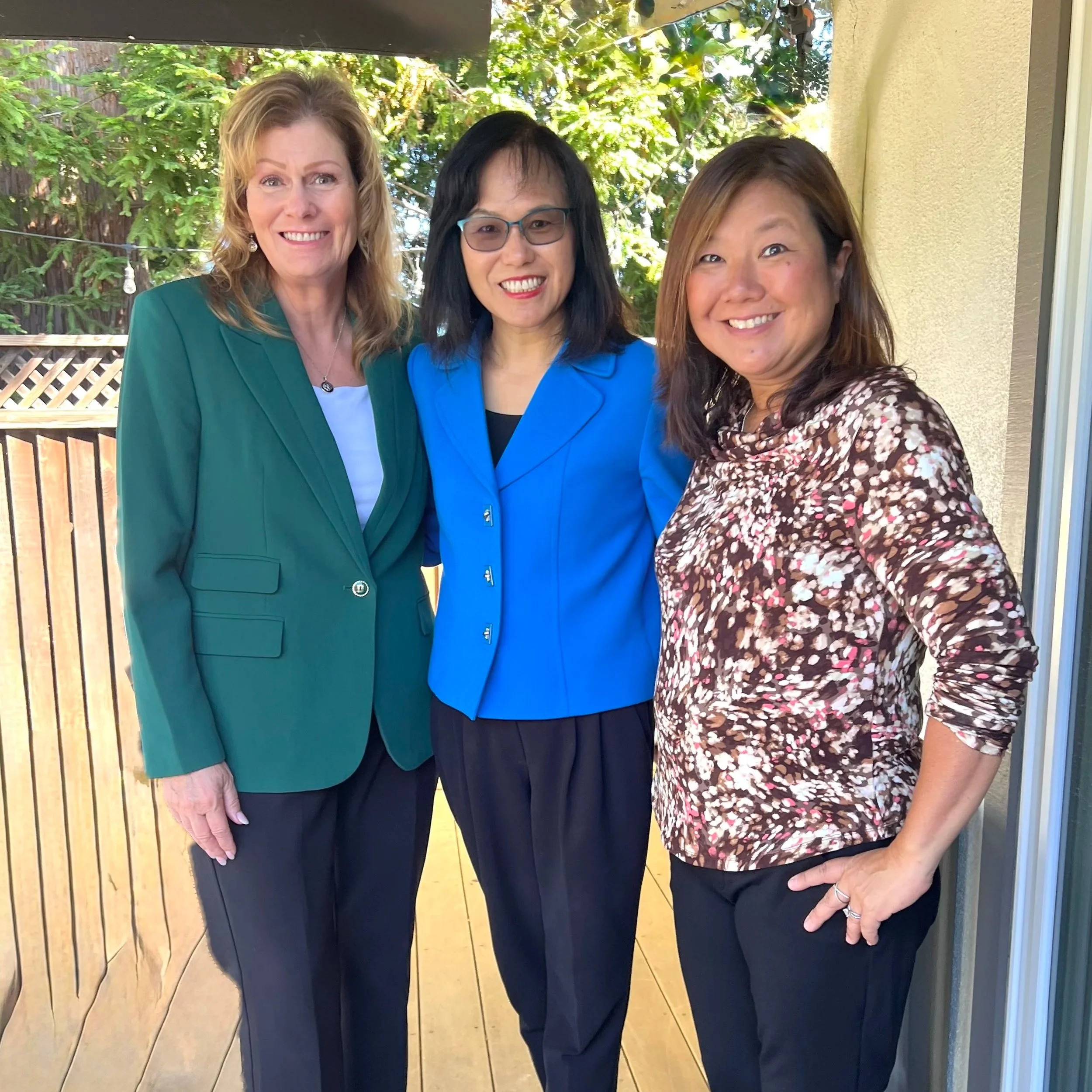 Three women smiling and taking a selfie outdoors. The woman on the left has blonde hair and is wearing a green blazer. The woman in the middle has black hair, glasses, and is wearing a blue blazer. The woman on the right has brown hair and is wearing