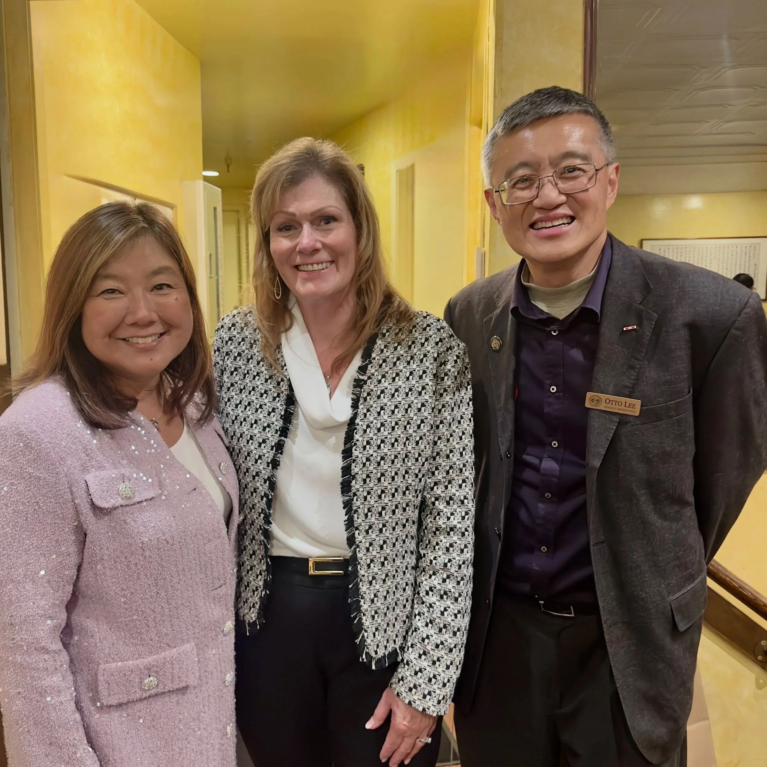 Group of three people standing together indoors, smiling for the camera. The woman on the left wears a pink coat, the woman in the middle has blonde hair and wears a patterned black and white jacket, and the man on the right has glasses, short gray h