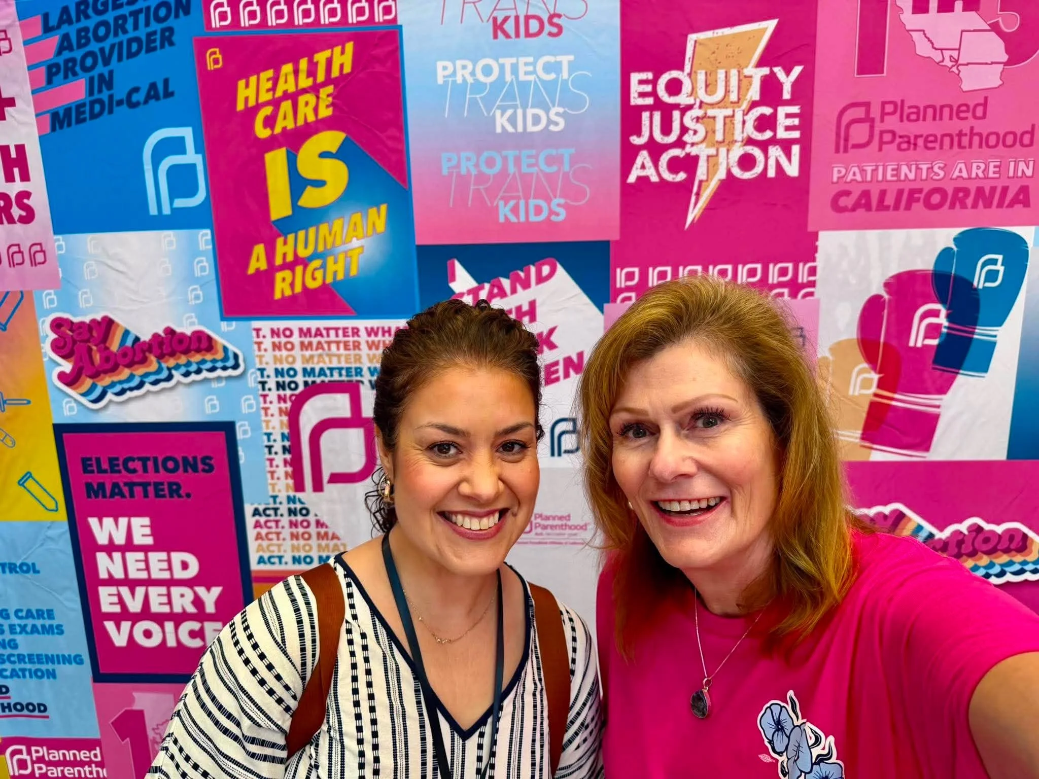 Two women smiling in front of a colorful background filled with signs and slogans related to Planned Parenthood and reproductive health issues, including messages about abortion rights, healthcare, and equity.