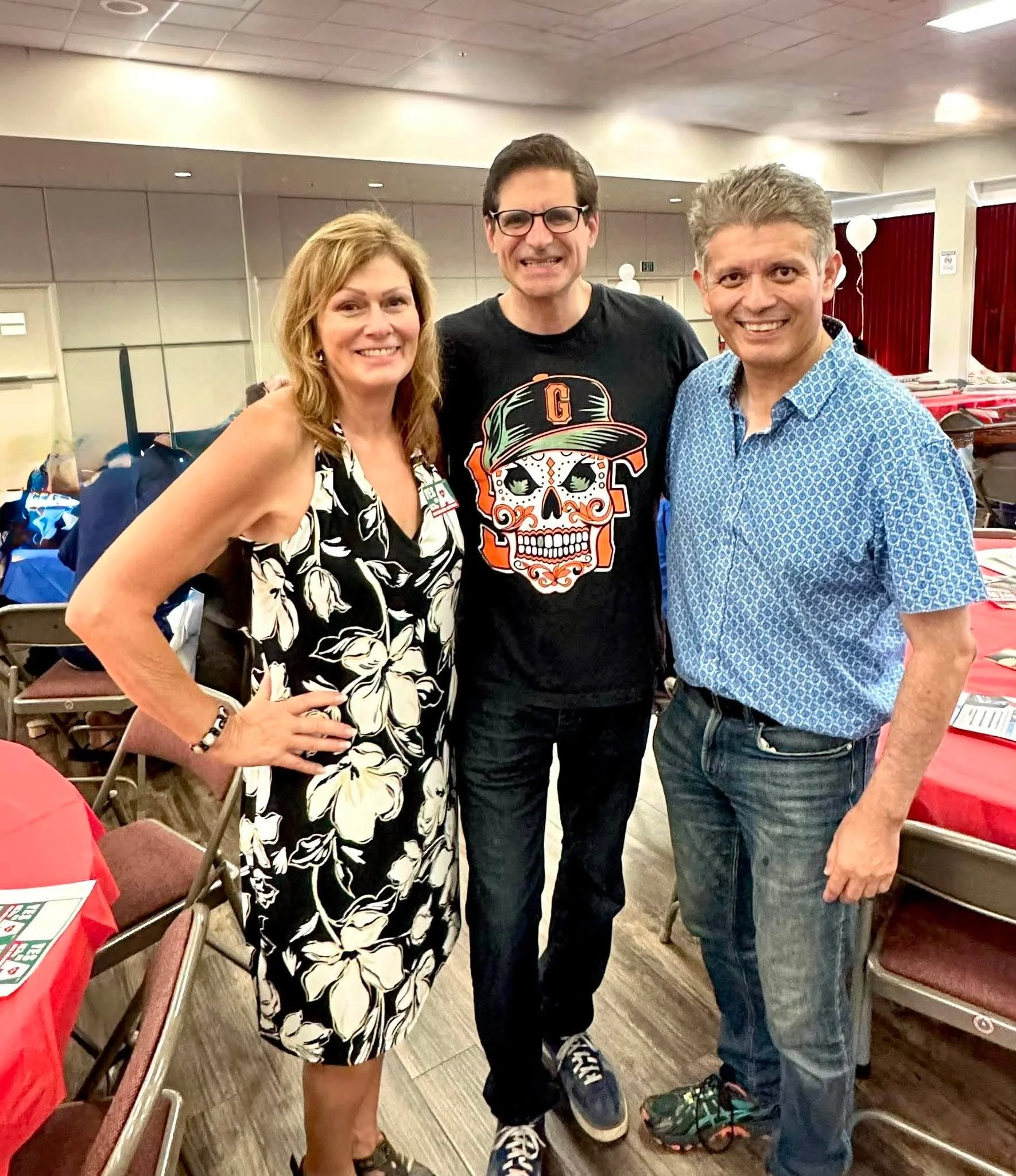 Three people standing close together smiling inside a room. The woman on the left is wearing a black and white floral dress, the man in the middle is wearing glasses and a black T-shirt with a colorful skull graphic, and the man on the right is weari