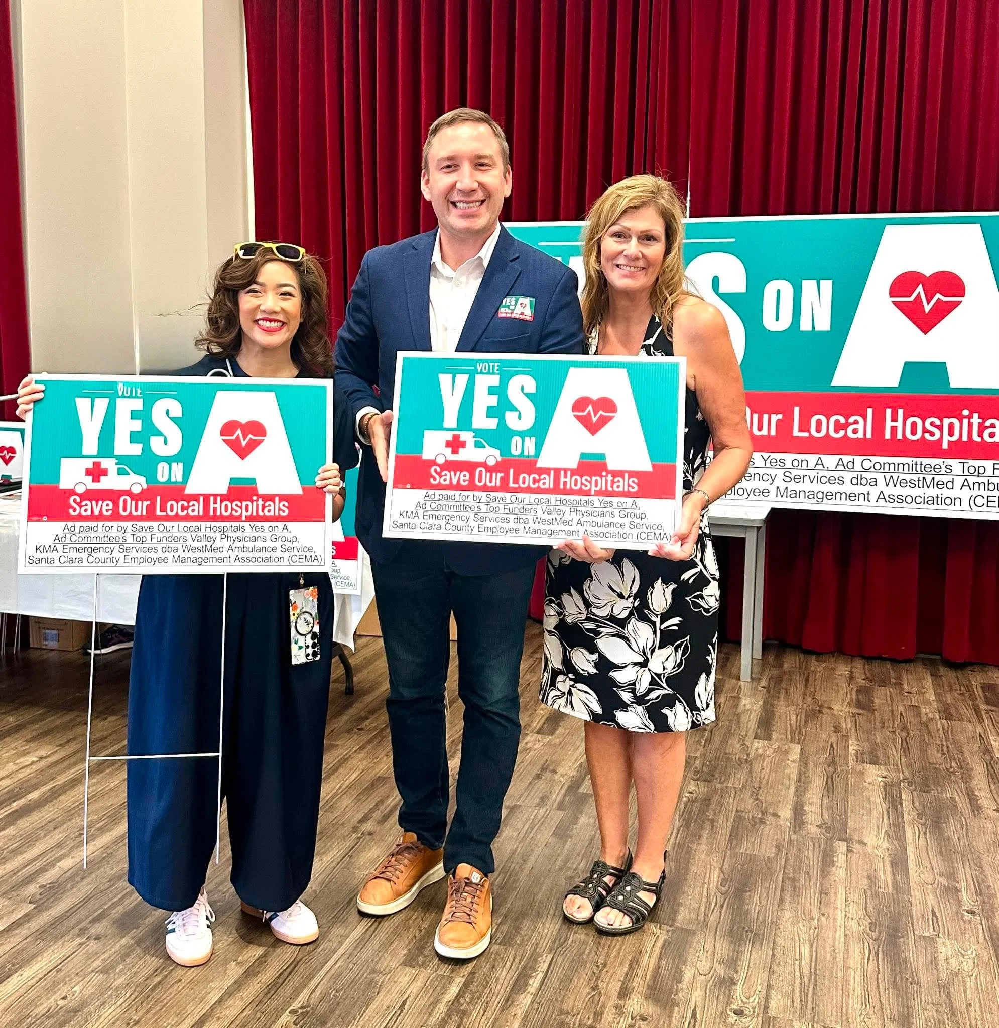 Three people holding signs at a health advocacy event, standing on a hardwood floor in front of red curtains. The signs promote saving local hospitals and include a heart symbol with a heartbeat line.