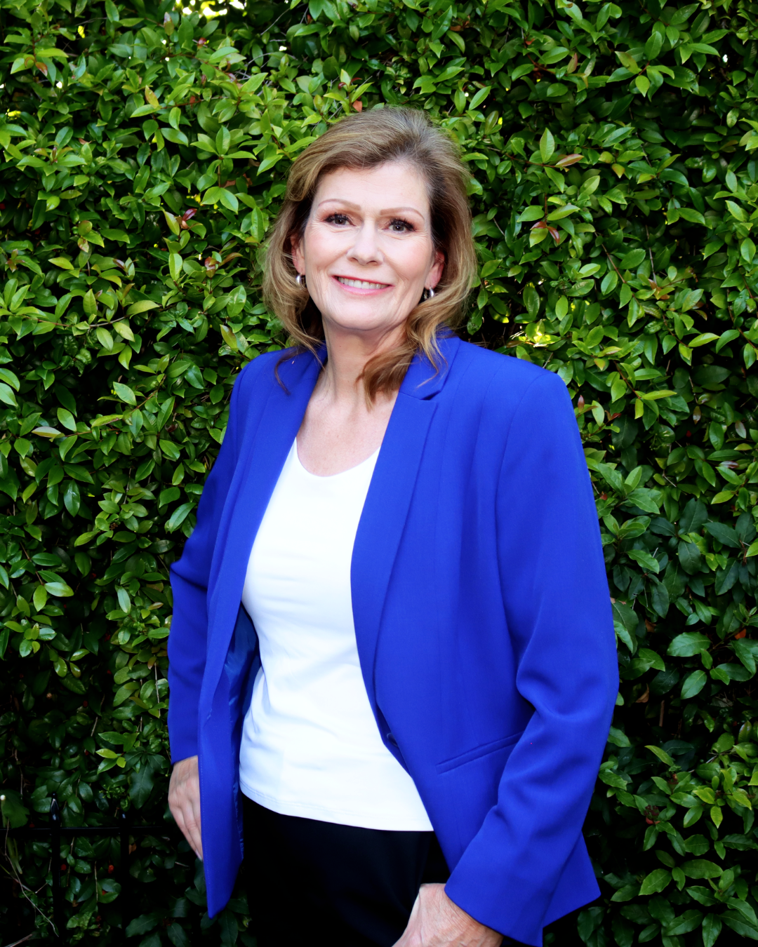 A woman with light brown hair in waves, wearing a royal blue blazer over a white top, standing in front of green leafy bushes, smiling at the camera.