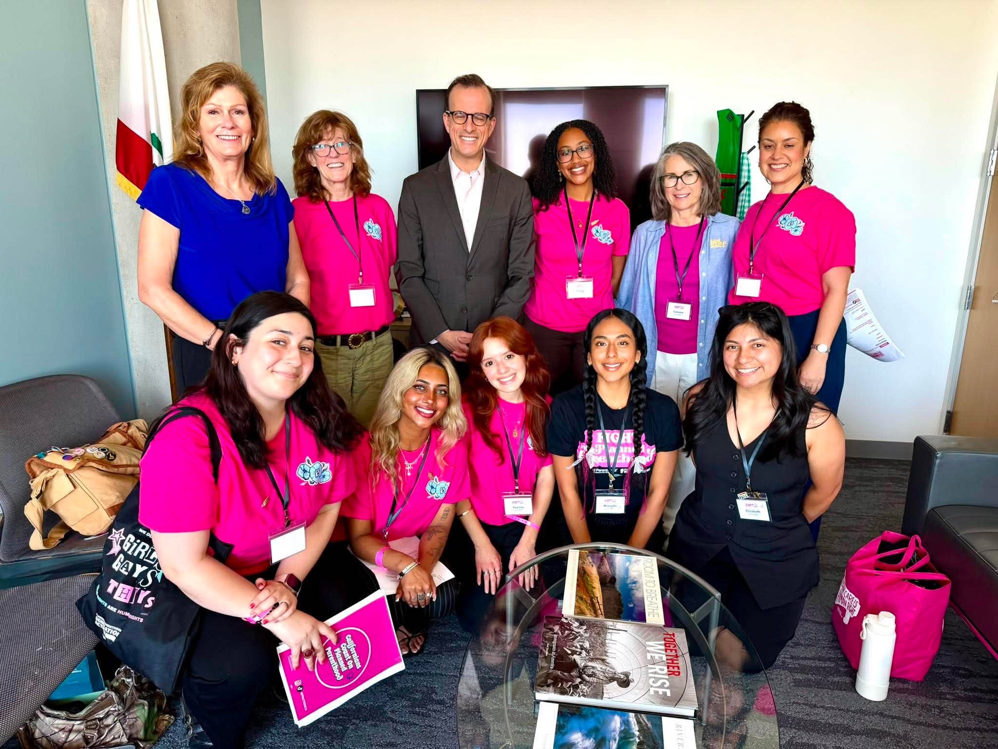 Group of diverse women and one man smiling and posing for a photo in a room, with some wearing pink shirts and badges, and others wearing casual clothing. Several women are sitting while others are standing behind them.