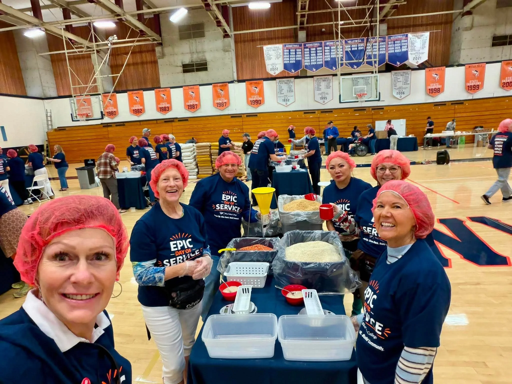 Group of people wearing pink hair nets and dark blue T-shirts with a logo, gathered in a gymnasium with banners on the walls. Some are smiling at the camera while others are working at tables with food and supplies. The gym has high ceilings, basketb