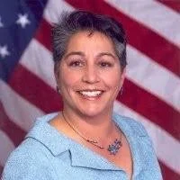 A woman smiling in front of an American flag background, wearing a light blue blazer and a necklace.