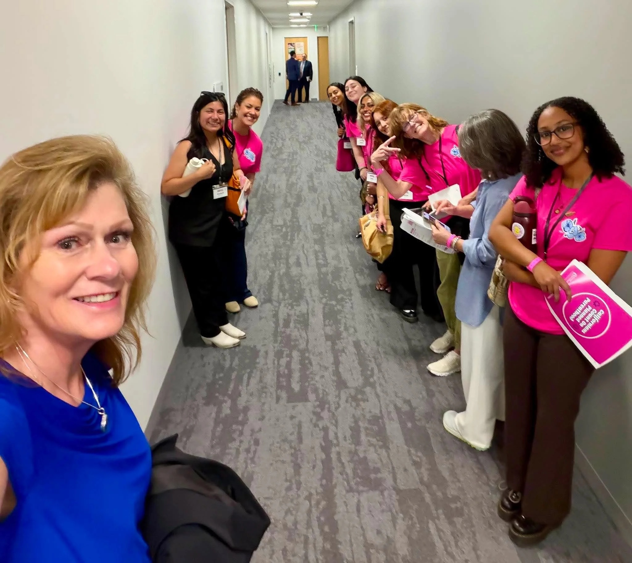 A group of women standing in a line along the wall in a hallway, all wearing pink shirts with various accessories and name tags, smiling and posing for the camera, with two men in suits talking in the background.