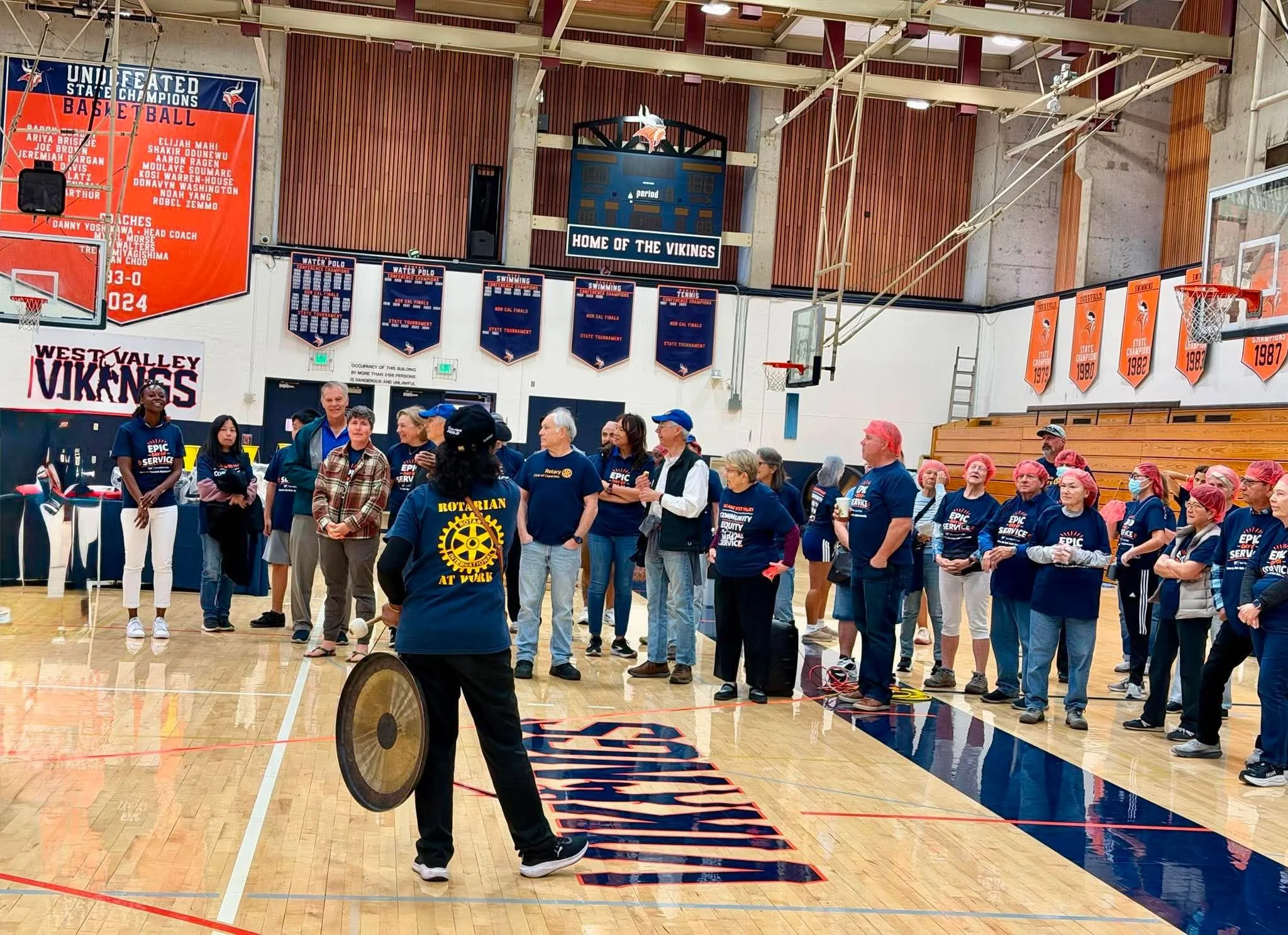 People gathered in a gymnasium for a community event, with banners celebrating the team and various sports activities on the walls.