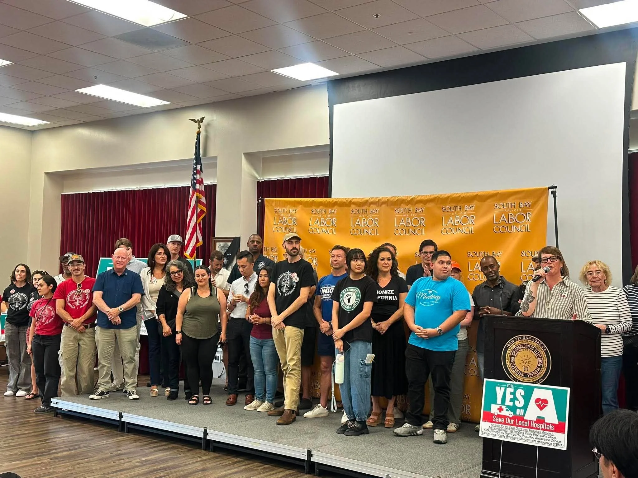 A diverse group of people gathered on a stage at a community event, with a yellow banner reading 'South Bay Labor Council.' Several individuals are standing and sitting in the front, and some are speaking into microphones. An American flag is in the 