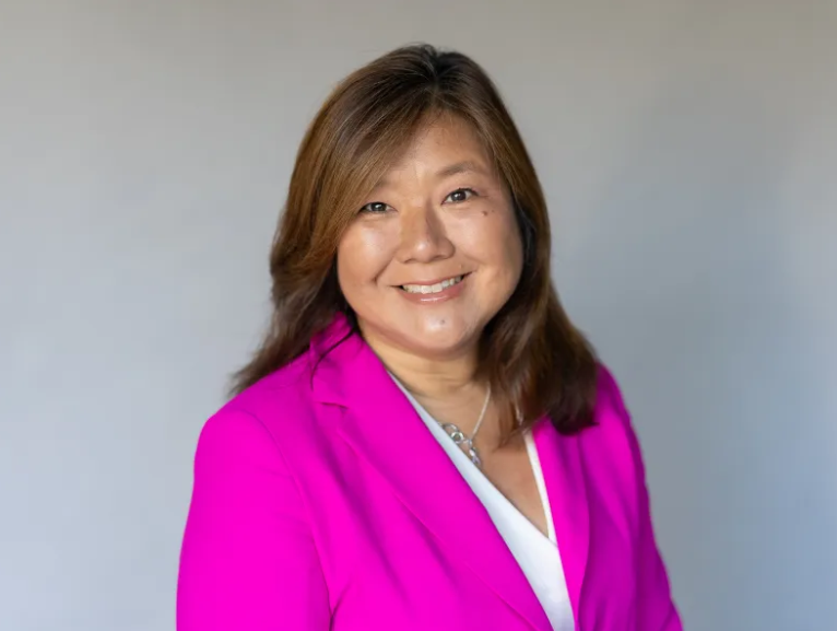 A woman with shoulder-length brown hair wearing a bright pink blazer and a white top, smiling at the camera.