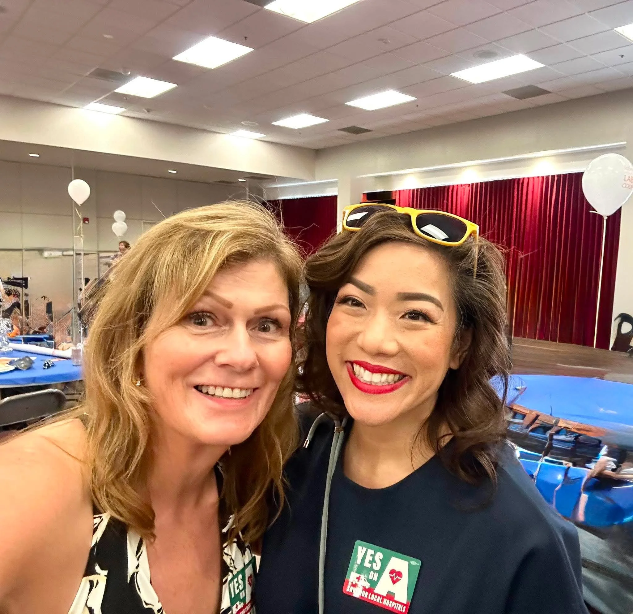 Two smiling women taking a selfie at an indoor event with balloon decorations and people in the background. One woman has blonde hair and the other has brown hair, both wearing black tops and 'YES' stickers for a health-related cause.