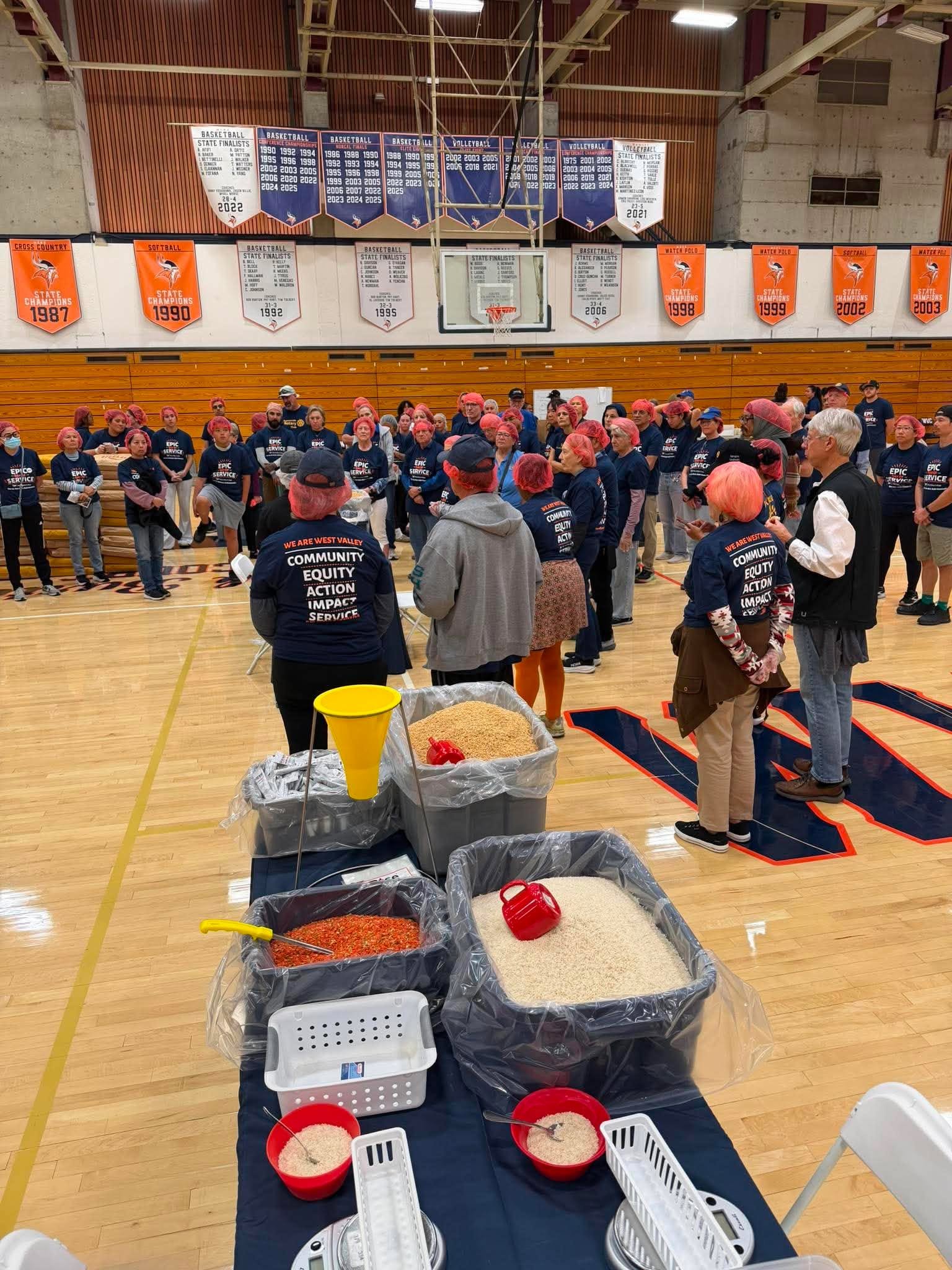 Group of people gathered inside a gymnasium, participating in a community service event focused on food collection and distribution. In the foreground, there are containers filled with rice, beans, and other grains, along with scoops and measuring cu