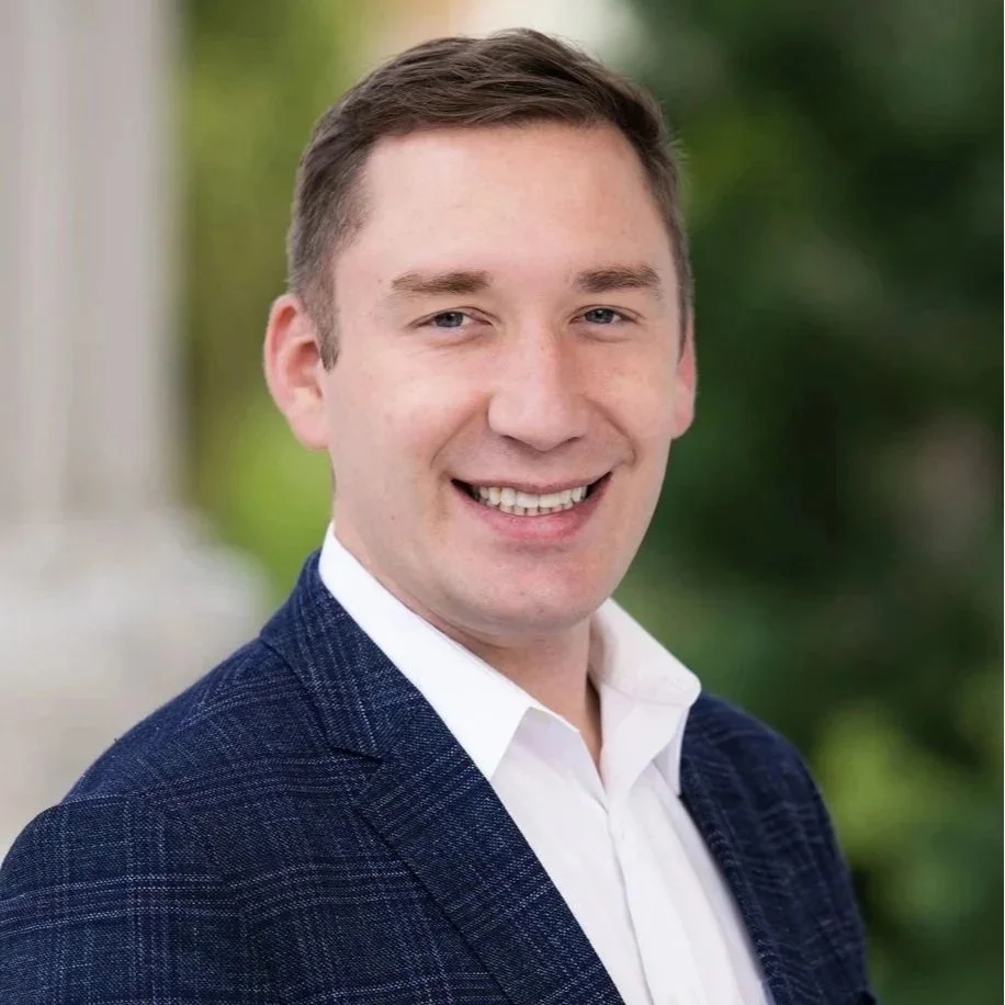 Patrick Ahrens in a white shirt and navy blazer smiling outdoors with blurred greenery in the background.