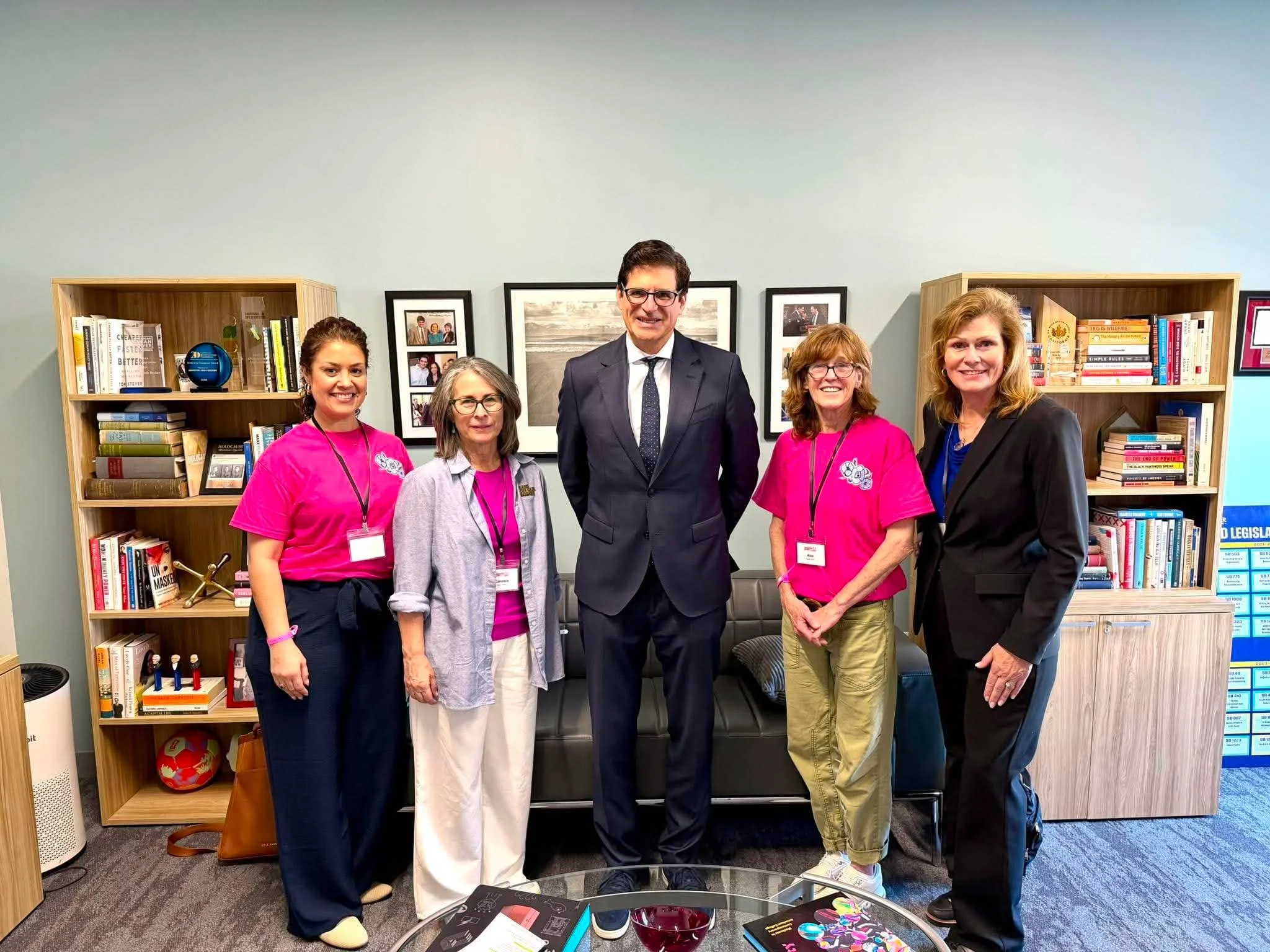 Group of five diverse women and a man standing together in an office or library, smiling at the camera. Two women are wearing pink shirts, and the man is in a dark suit with glasses. The background has bookshelves filled with books and framed photogr