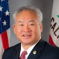 Portrait of Paul Fong with gray hair, wearing a dark suit, white shirt, and red tie, standing in front of the American flag and a California flag.