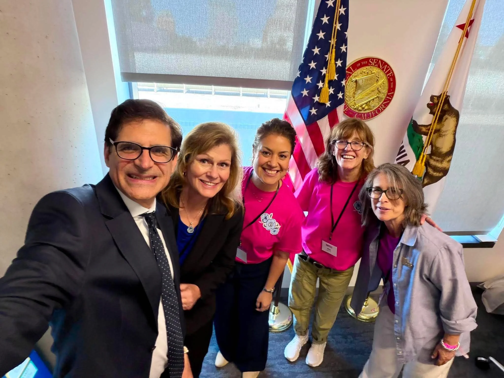 Group of people taking a selfie in front of flags, including the United States flag and California state flag, at an indoor event.