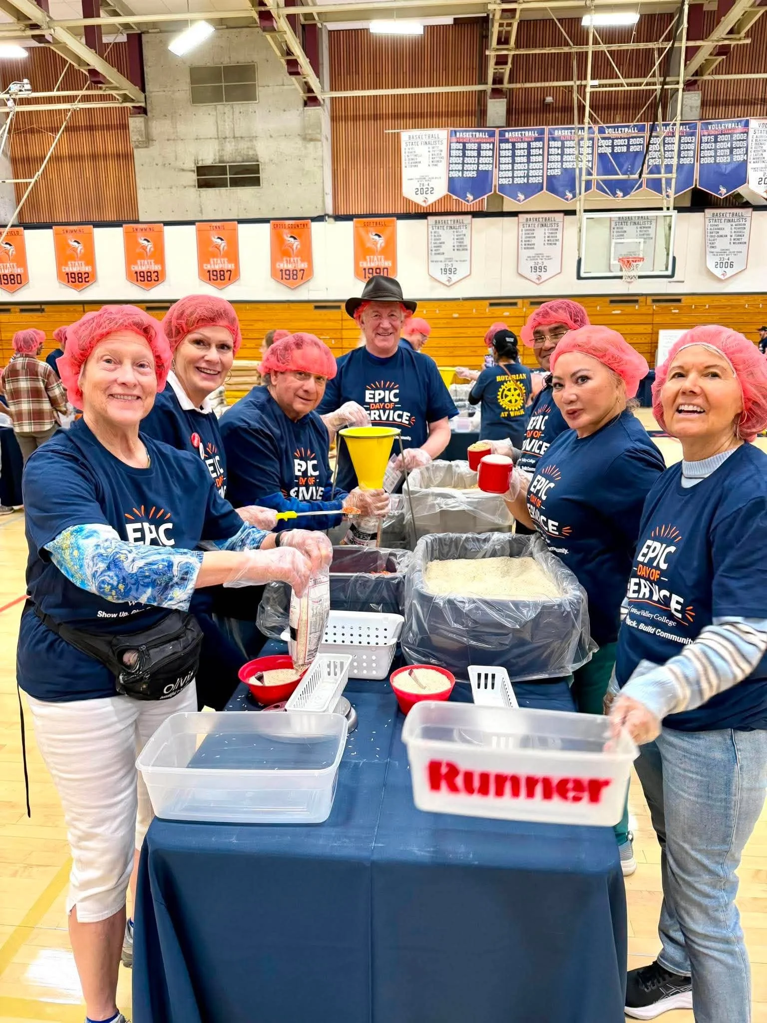 A group of people in a gymnasium participating in a food packaging event. They are wearing blue shirts with the text 'EPIC Day of Service' and pink hairnets. There are large containers of rice on the table, and banners with athletic achievements hang