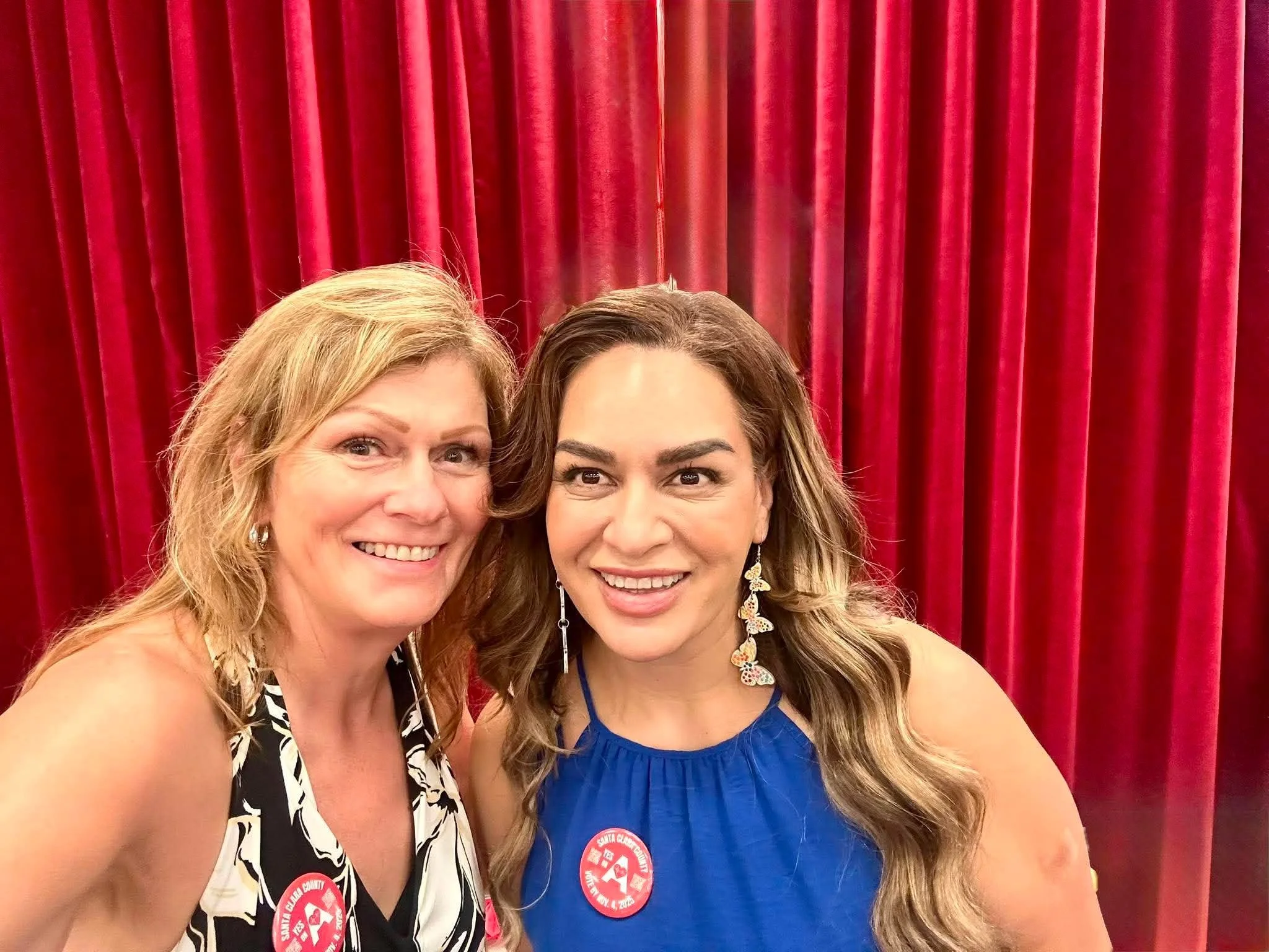 Two women smiling in front of red curtain, both wearing badges related to Santa Clara County.