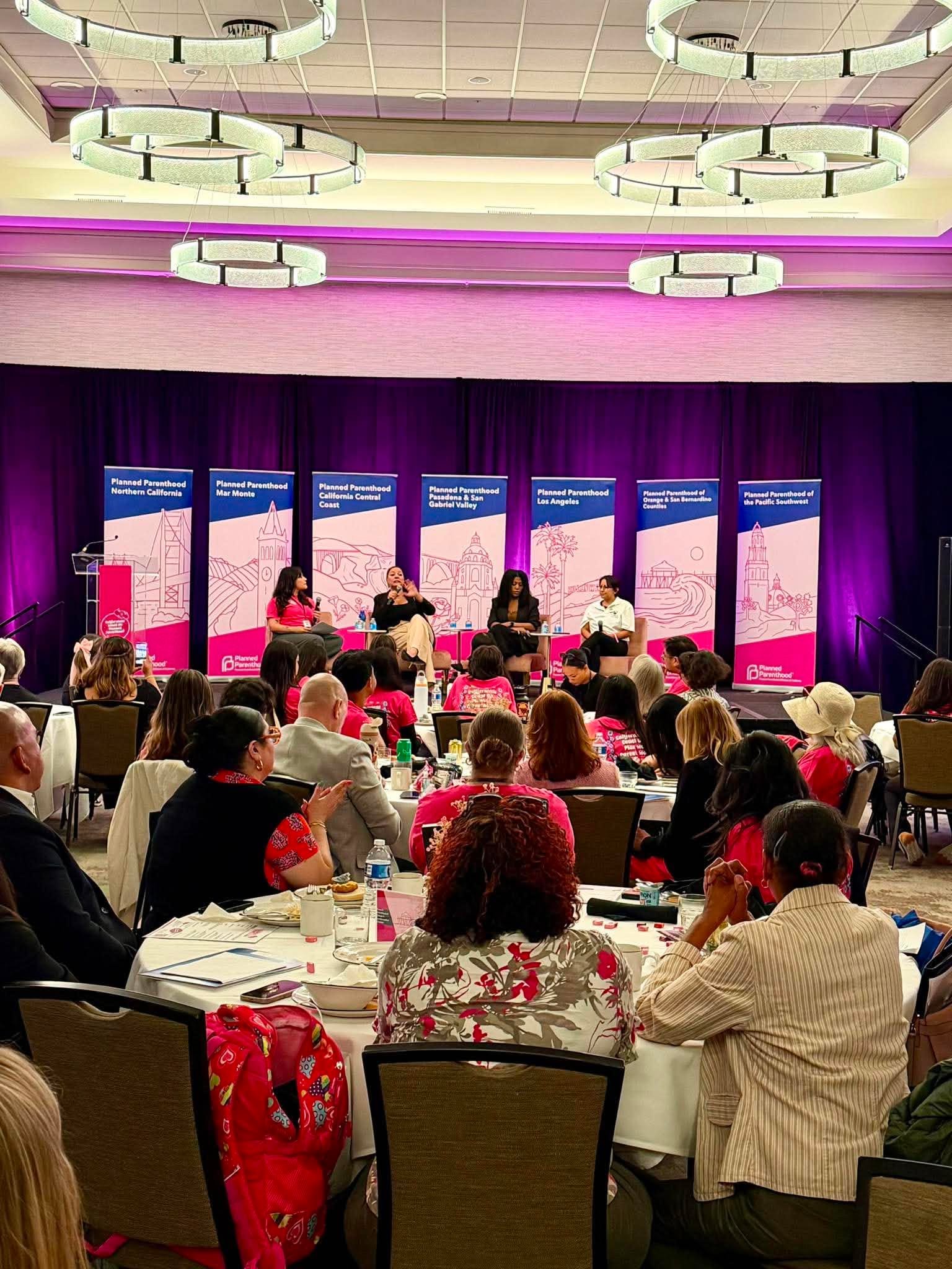 A panel discussion at a conference with four women speakers on stage and an audience seated at tables, some taking notes and photos. The backdrop displays banners related to Planned Parenthood.