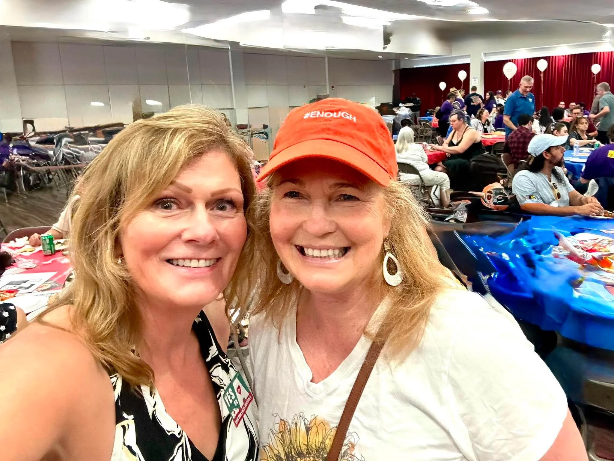 Two women smiling for a selfie at a crowded indoor event with tables, chairs, and people in the background.