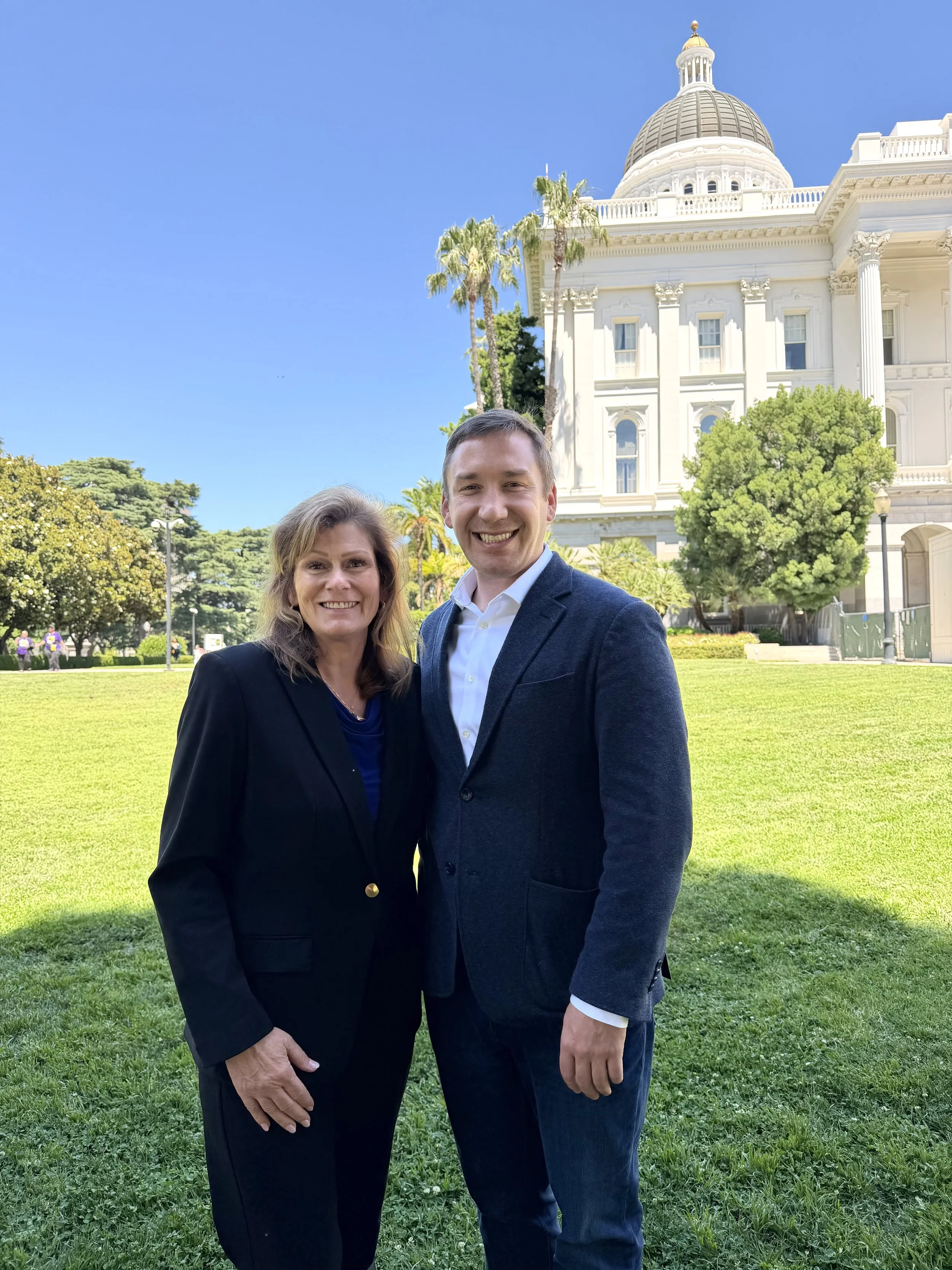 A woman and a man standing together outdoors in front of the California State Capitol building, smiling at the camera, with trees and a lawn around them against a clear blue sky.
