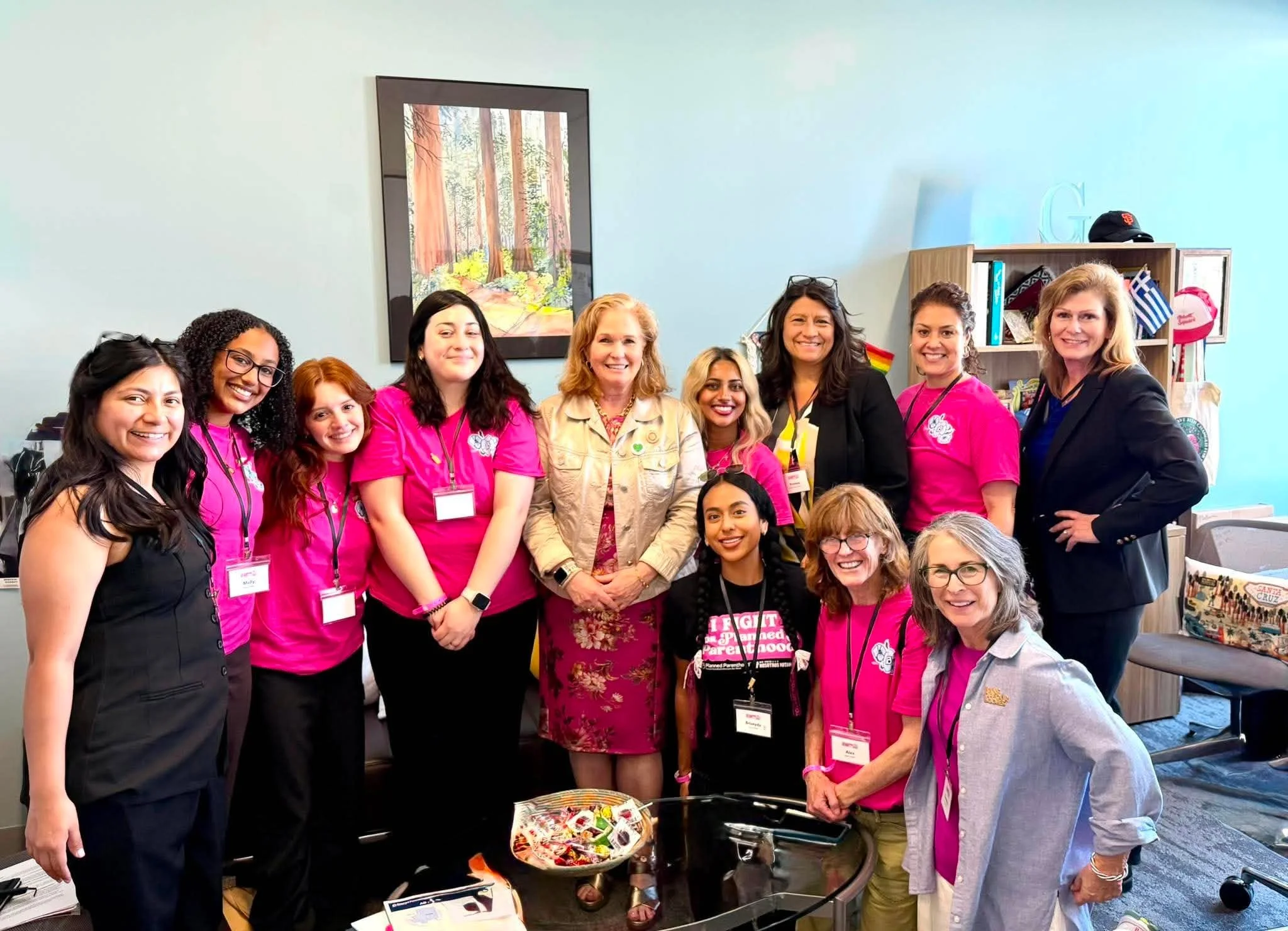 Group of women smiling and standing together at an indoor event, some wearing pink shirts and context-related badges, with books and flyers displayed in front.