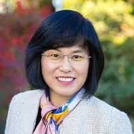 A woman with short black hair, wearing glasses, a light-colored blazer, and a colorful scarf, standing outdoors with trees in the background.
