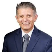 A professional man with short gray hair, wearing a navy blue suit, light blue shirt, and dark tie, smiling against a white background.