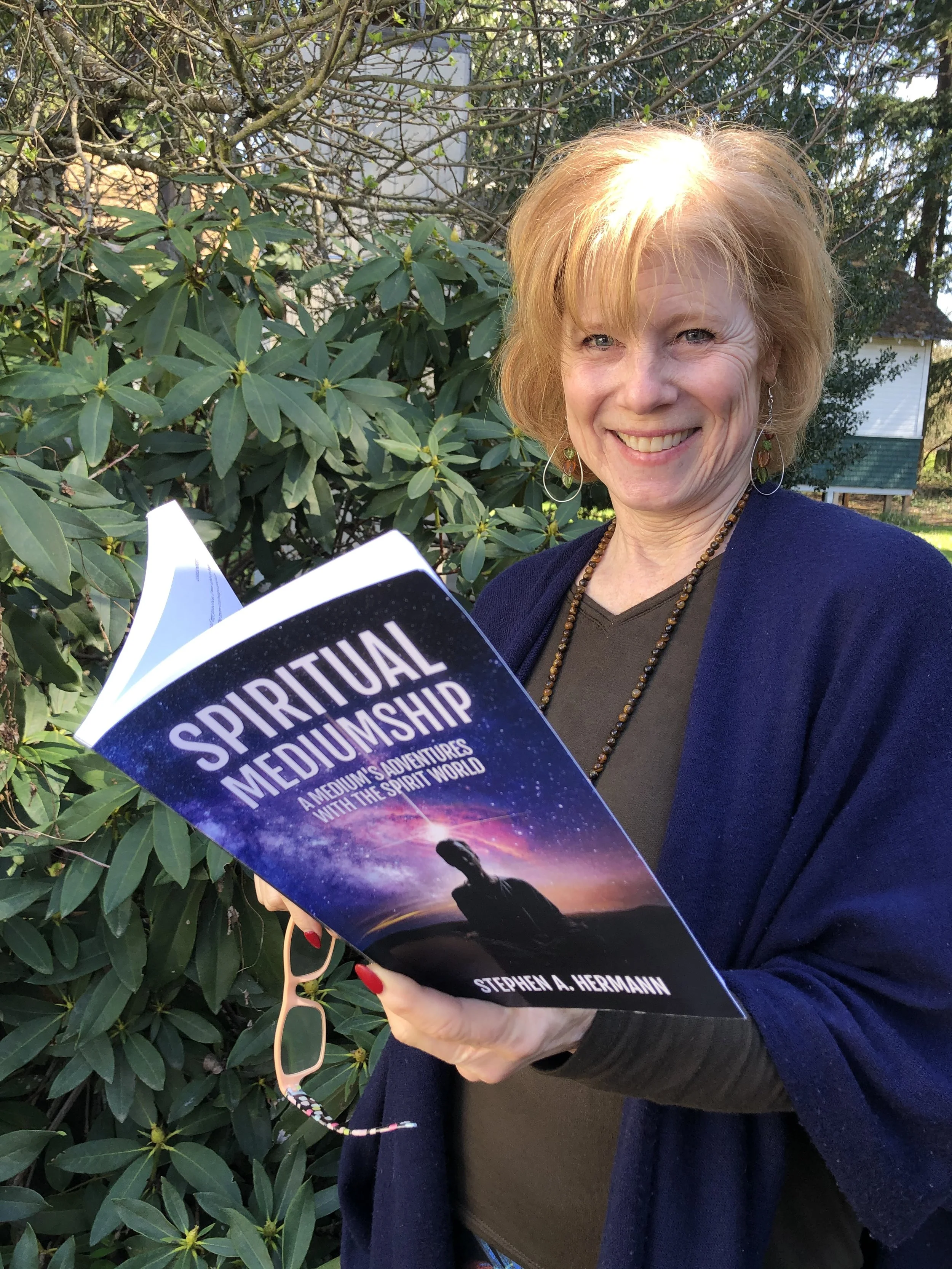 A woman with short blonde hair and earrings smiling outdoors, holding a book titled 'Spiritual Mediumship' by Stephen A. Hermann, with green bushes and a house in the background.