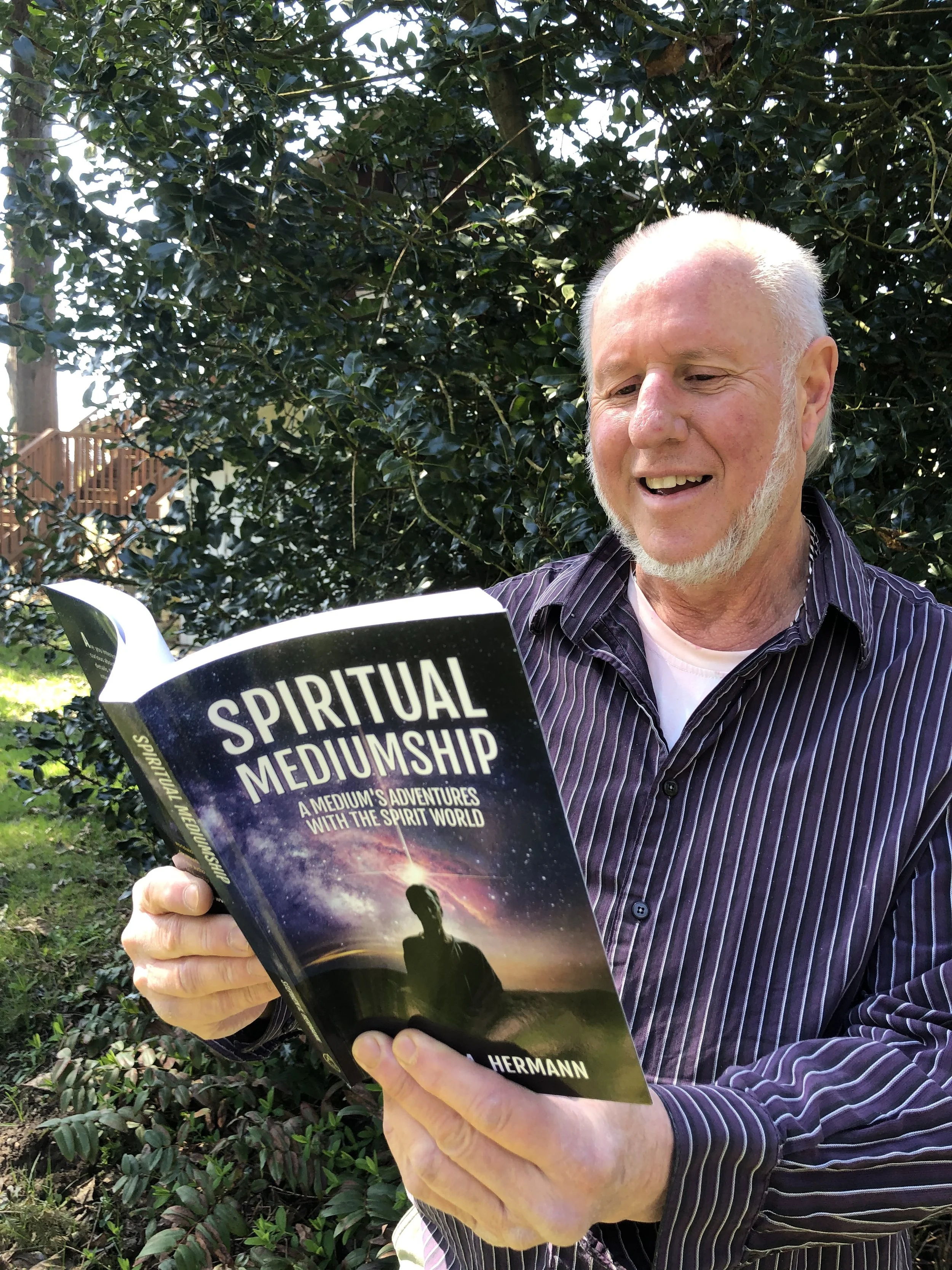 An older man with a white beard and striped shirt holding a book titled 'Spiritual Mediumship' outdoors, with green foliage in the background.