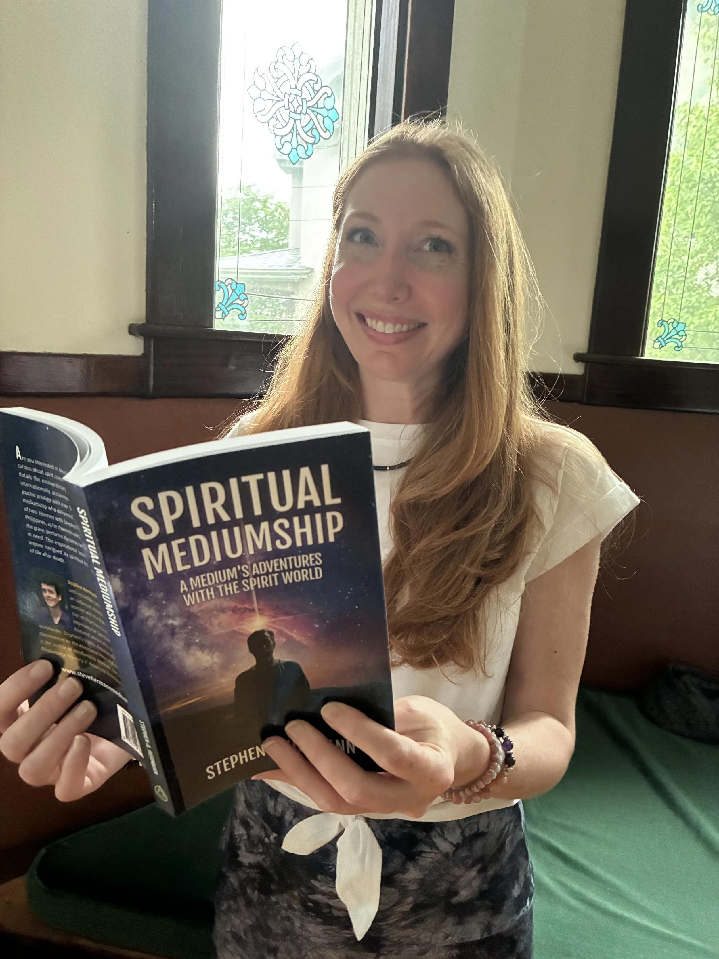 A woman with long red hair wearing a white blouse and a pearl bracelet, sitting in a room with stained glass window designs, reading a book titled 'Spiritual Mediumship'.