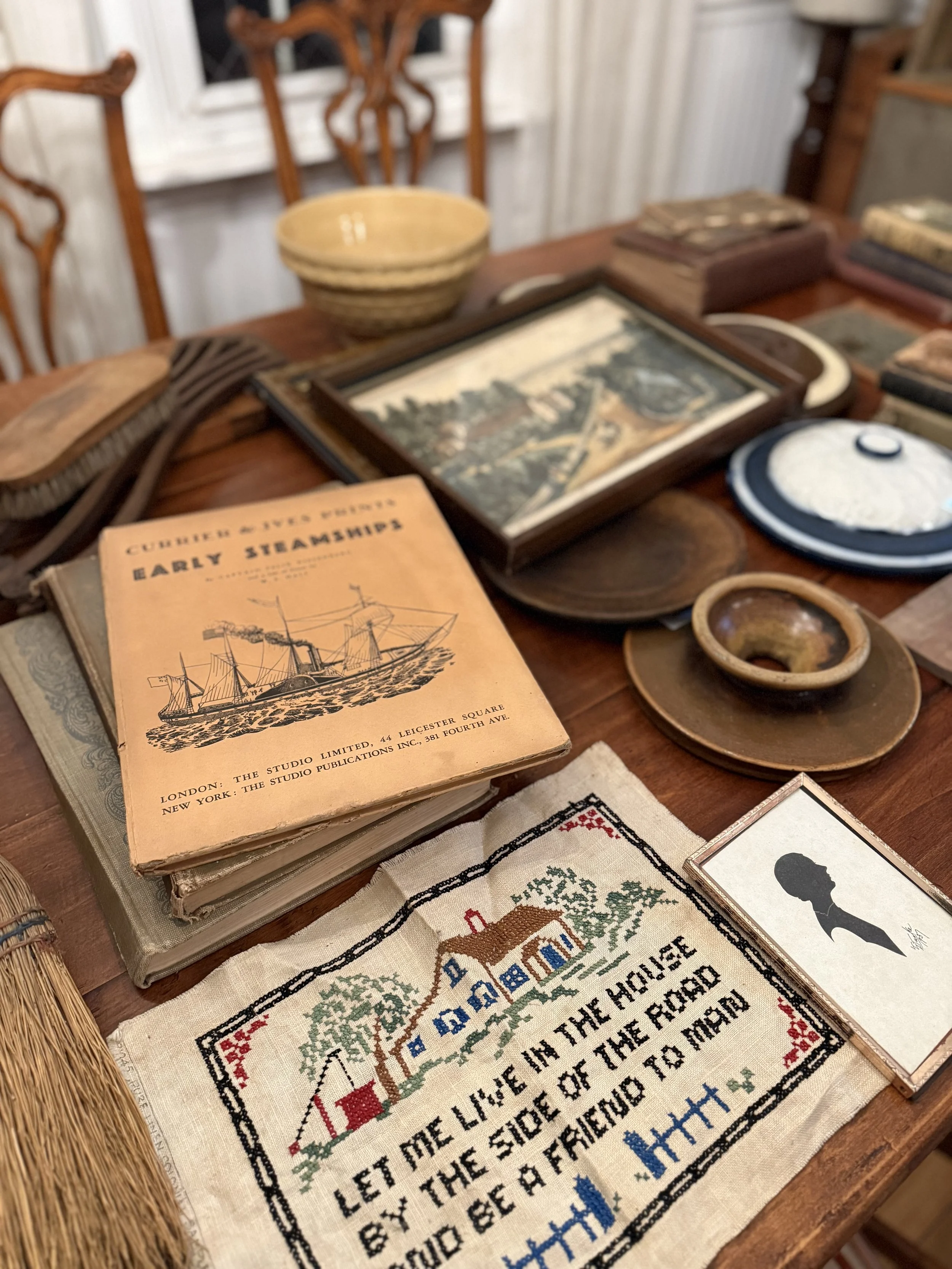 A wooden table displaying vintage books, framed pictures, pottery, and a cross-stitched fabric with a house silhouette and a poem, with wooden chairs in the background.