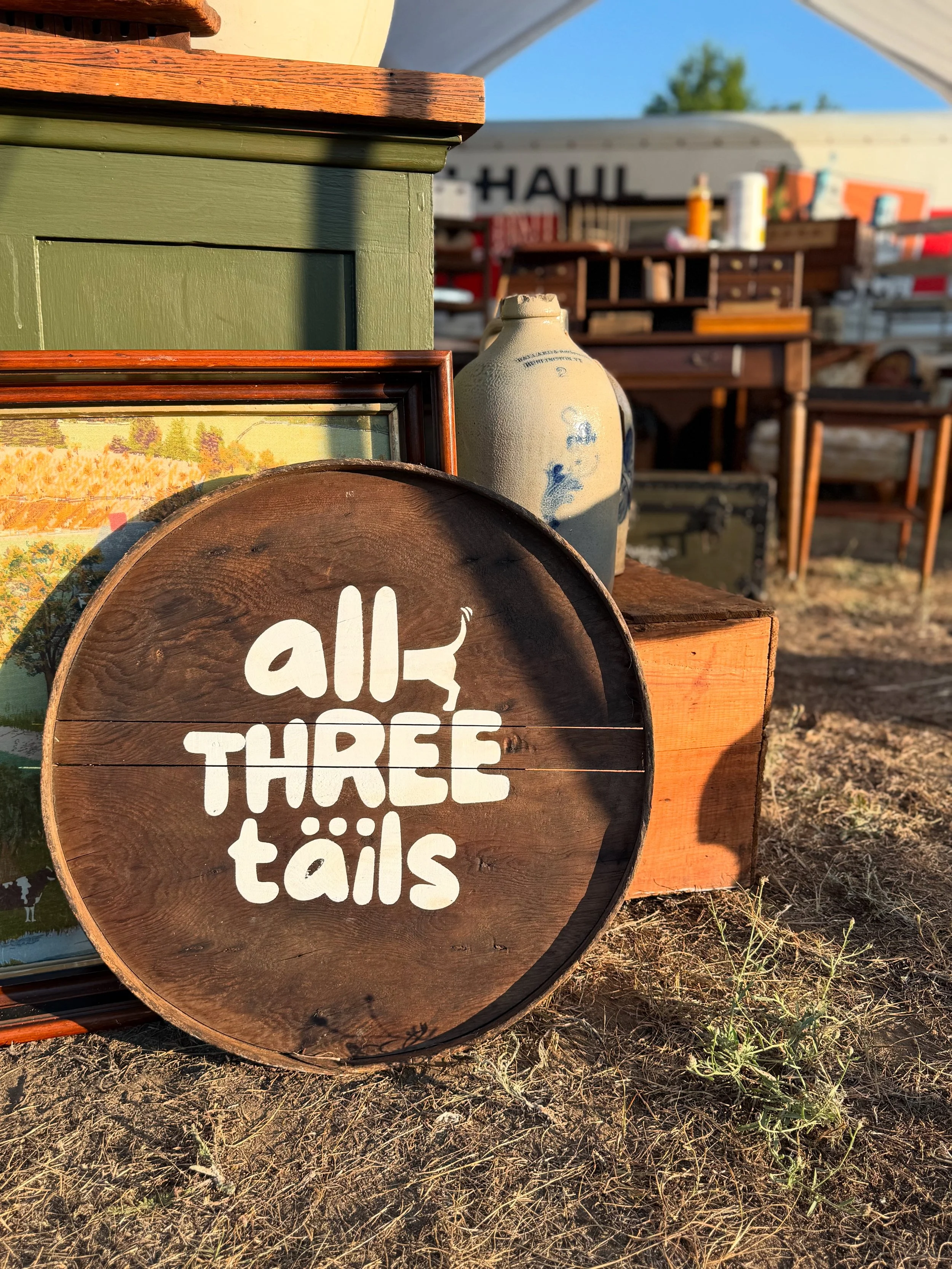A display table with framed artwork, a ceramic vase with blue floral patterns, and a round wooden sign that reads 'all three titles' in white text. The background shows a yard sale setup with boxes, furniture, and a U-Haul truck.