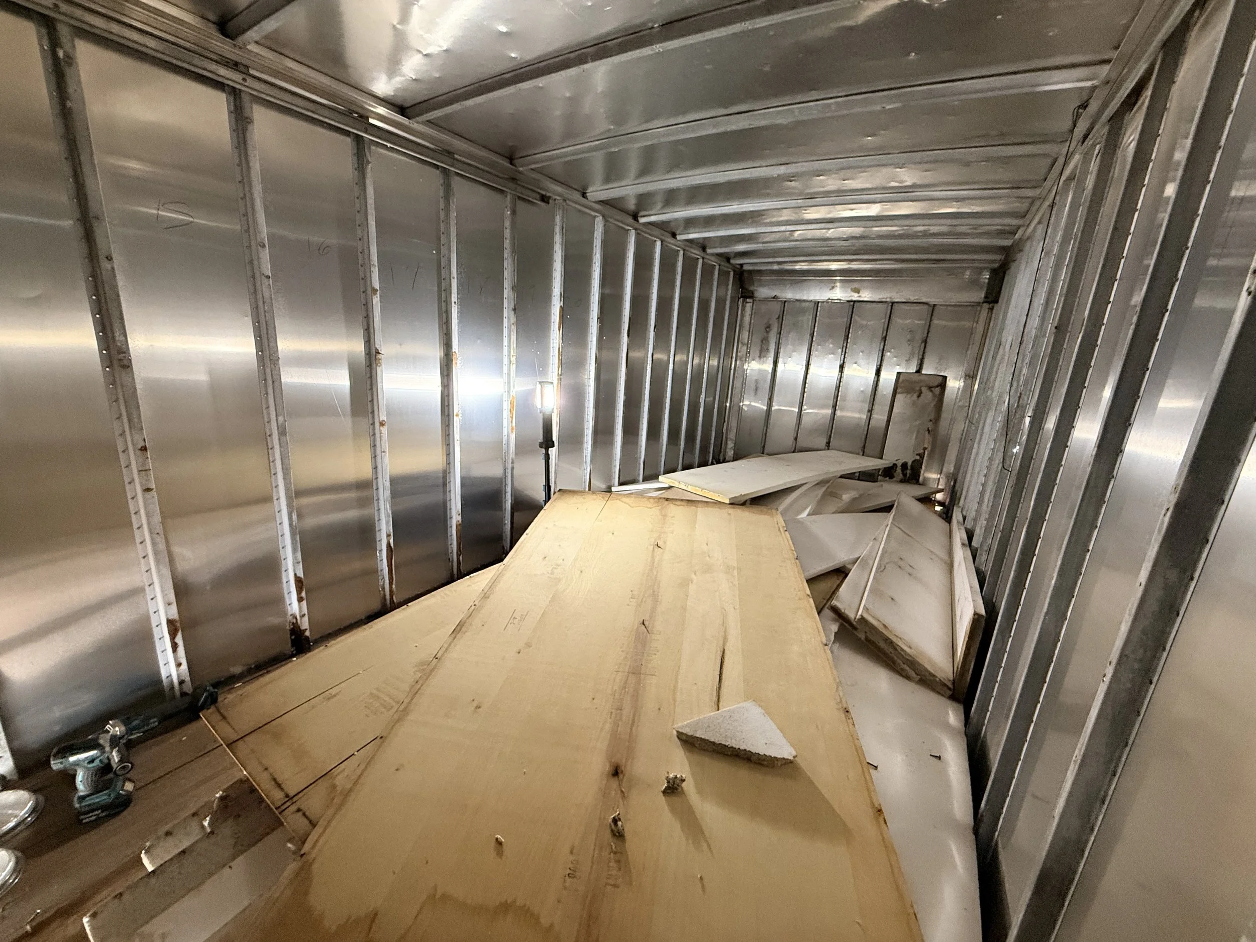The interior of an unfinished metal room or storage container with exposed metal framing and plywood flooring, construction debris, and a small light fixture.