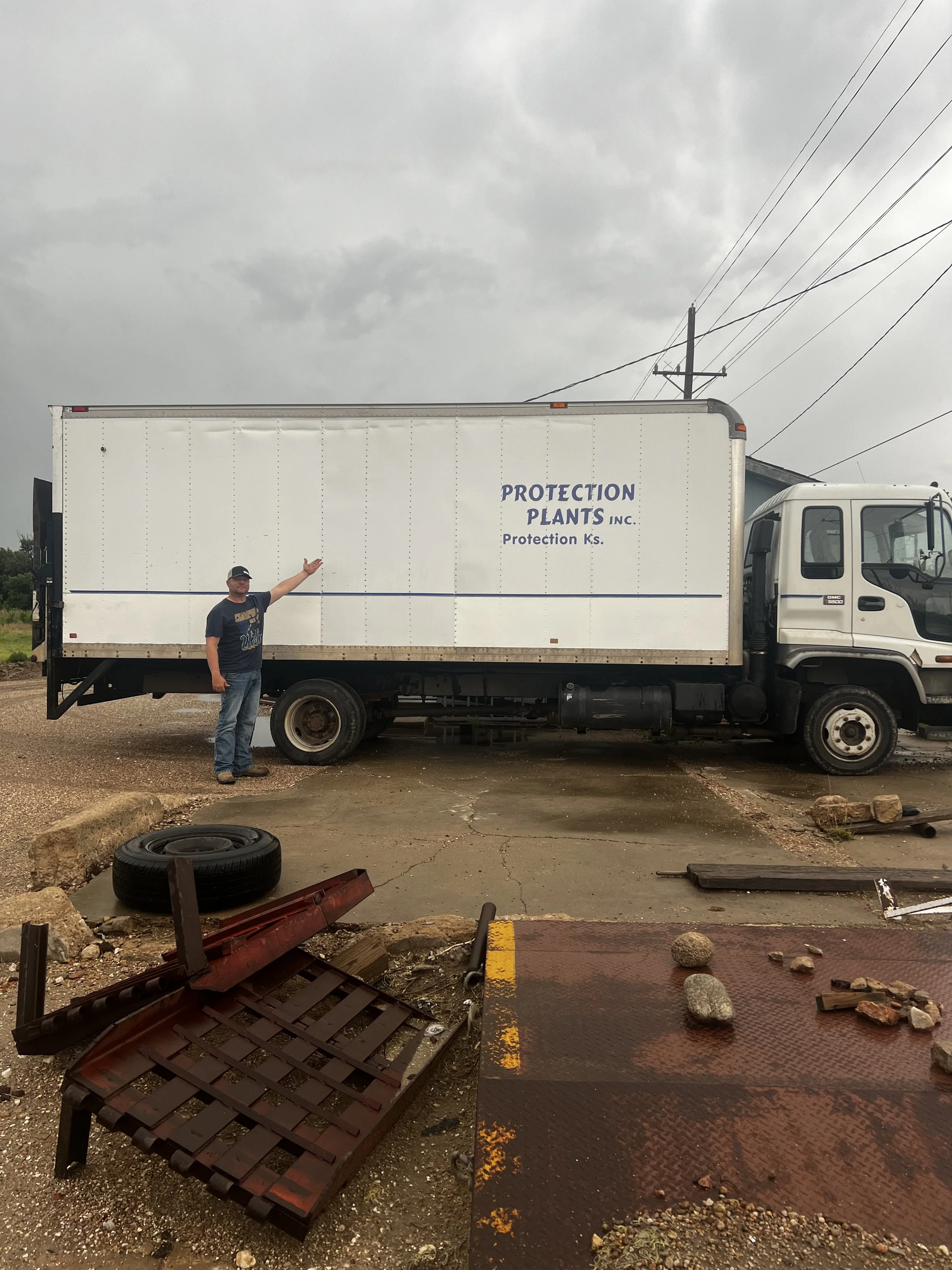 A man is standing outside next to a large white truck with the words 'Protection Plants Inc.' printed on the side. The man is pointing at the truck. The sky is overcast. The ground shows some debris, rocks, and a fallen sign or frame.
