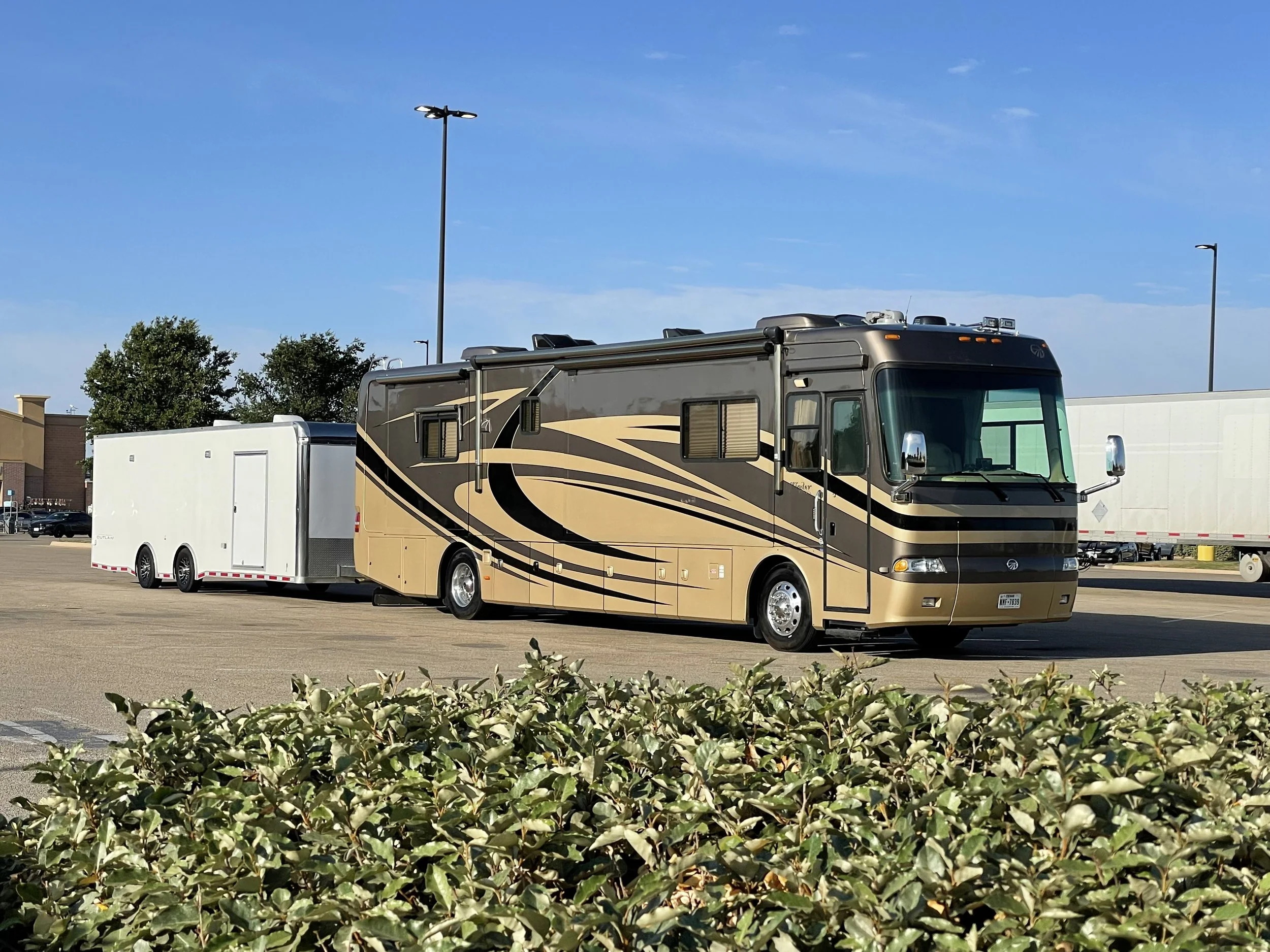 A large motorhome with brown and tan stripes is parked in a lot with a white trailer attached. The background features a blue sky with a few scattered clouds, trees, and a commercial building. In the foreground, there are bushes with green and white 