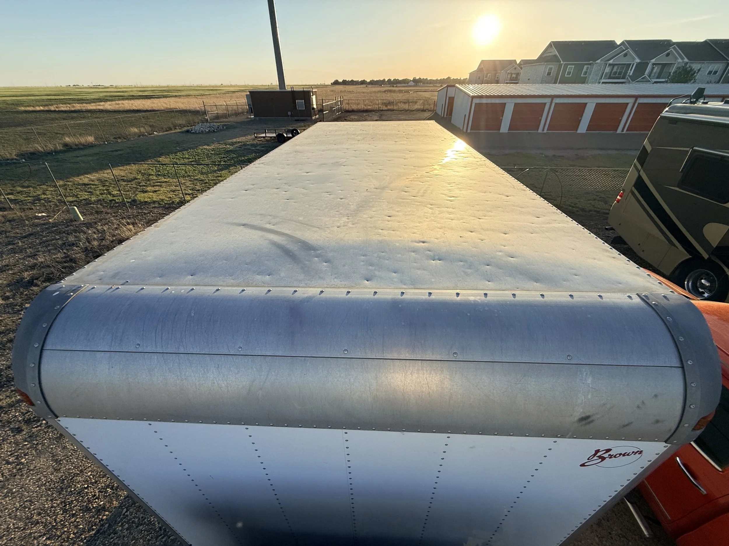 The image shows a close-up view of a portable metal trailer or camper with a reflective surface, in an outdoor area at sunset. In the background, there are residential houses and an open field.