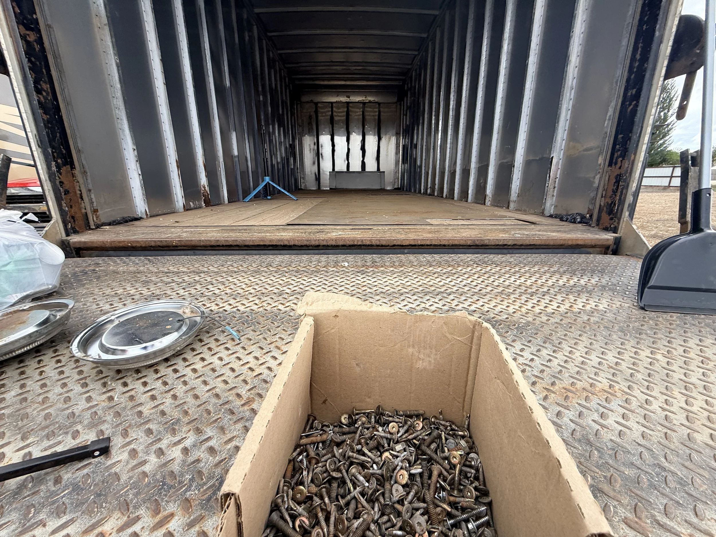 Inside of an empty truck trailer with metal walls, wooden floor, and scattered parts including screws, metal lids, and a cardboard box filled with more screws at the foreground.
