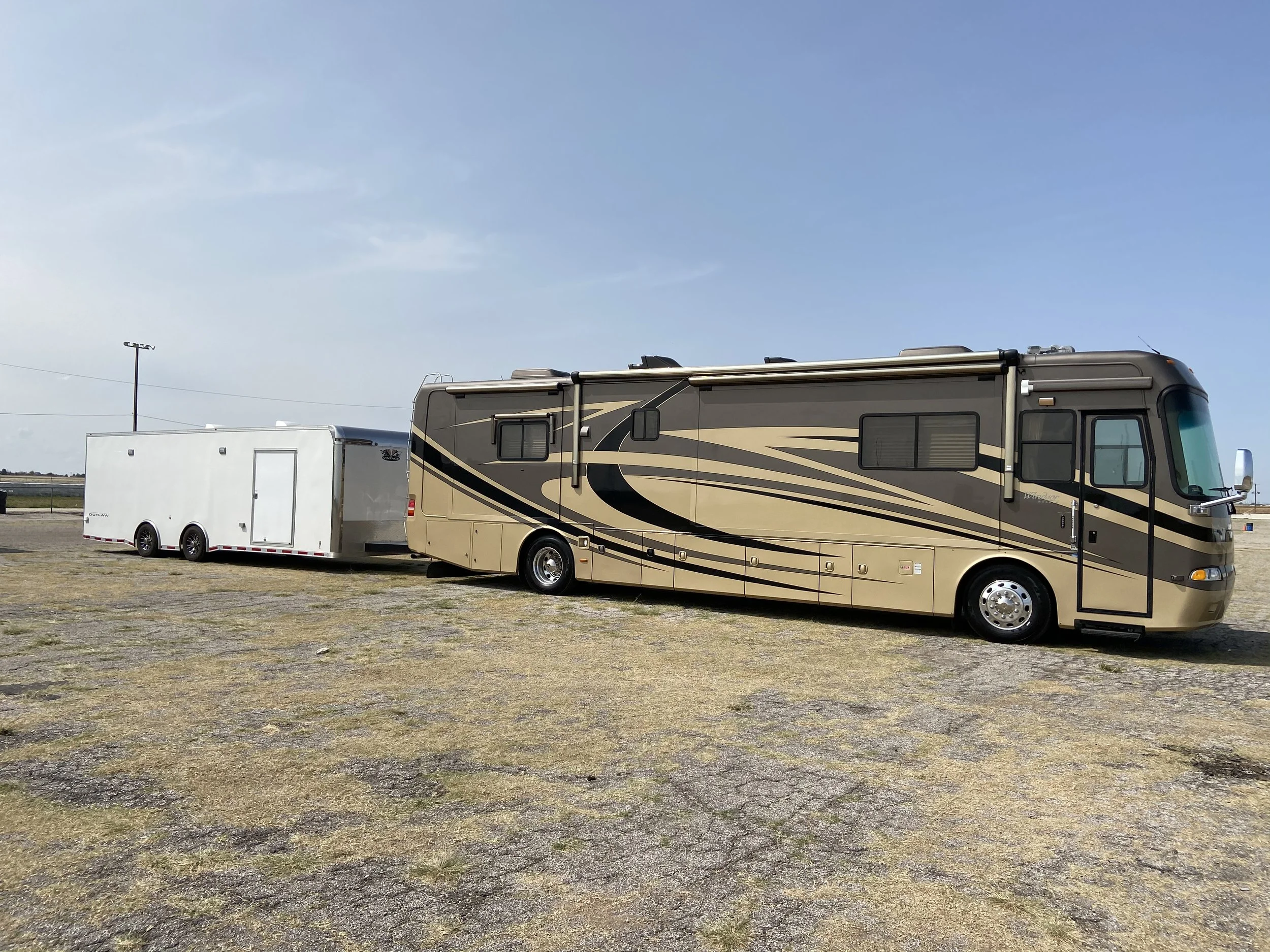 A large motorhome with a brown and beige swirl pattern parked on a patchy, grassy area, with a white trailer attached, under a clear blue sky.
